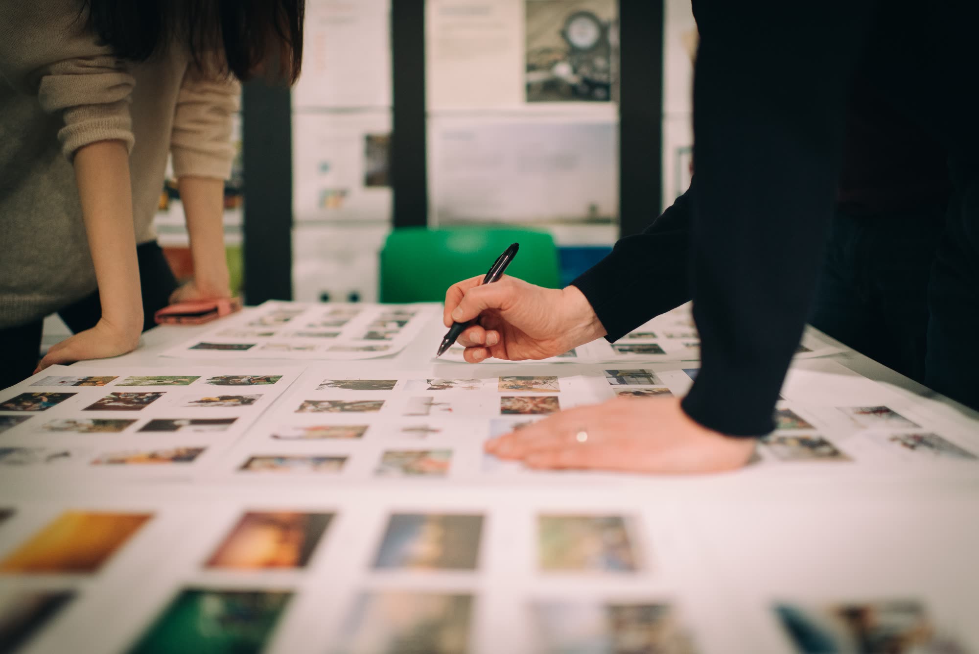 Two people leaning over a table covered with printed photo contact sheets, one marking a sheet with a black pen