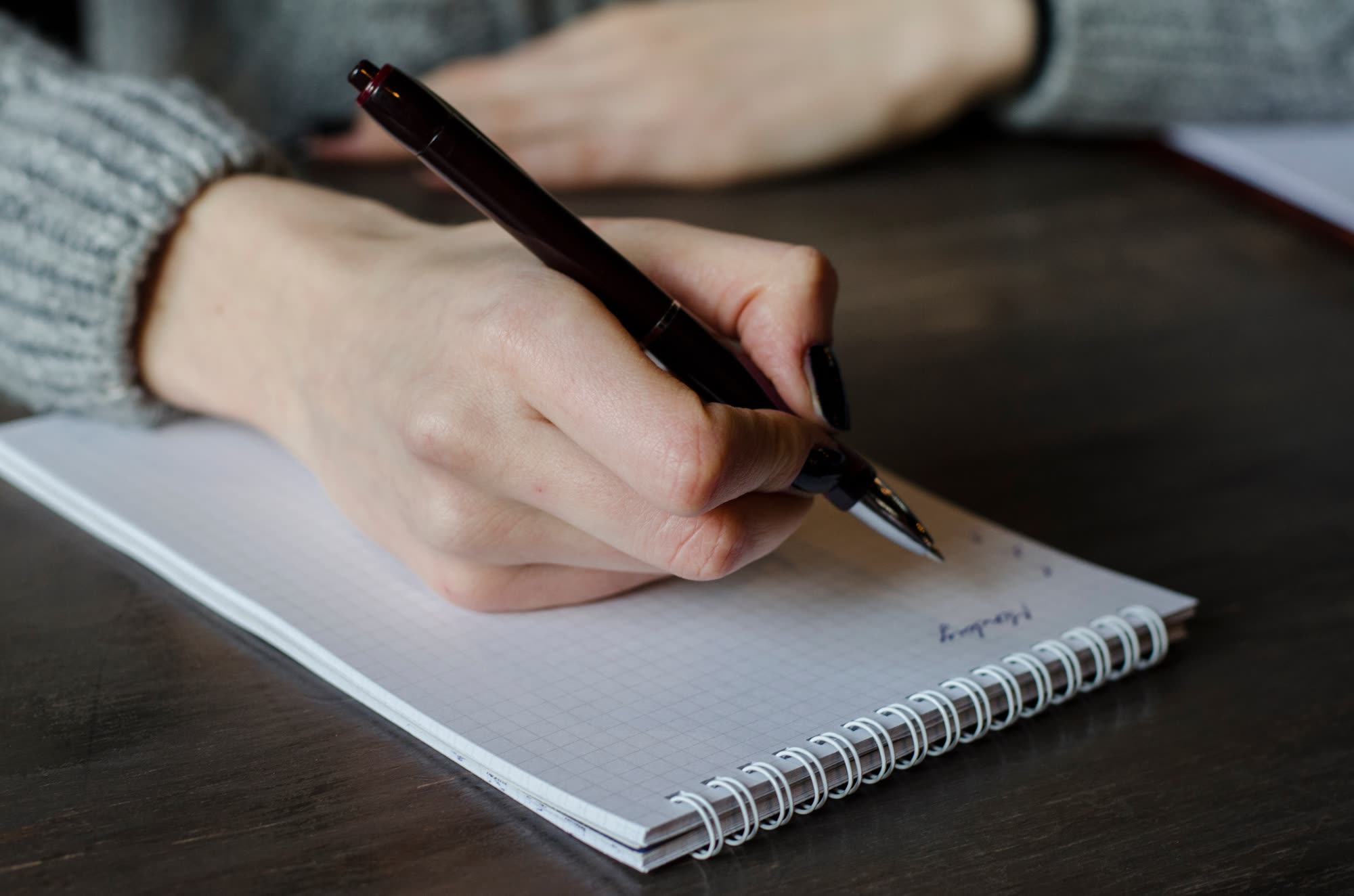 A hand writing in a spiral-bound squared notepad with a dark ballpoint pen on a wooden desk