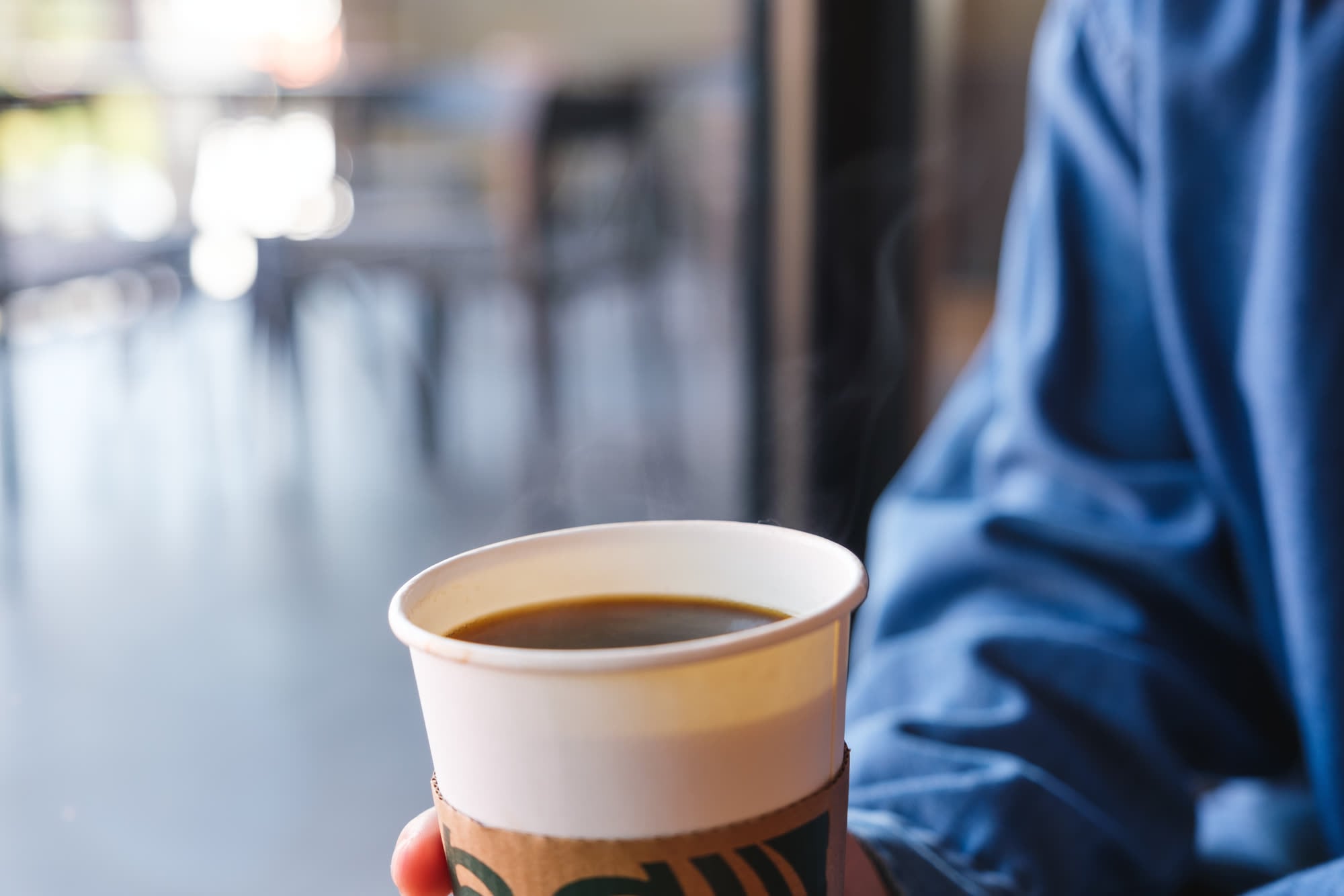 A hand holding a takeaway cup of black coffee in a cardboard sleeve, steam rising, a café interior blurred behind