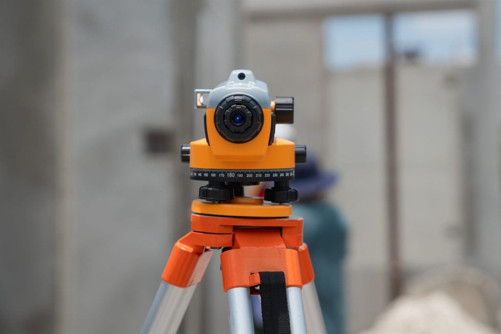 An orange surveyor's level mounted on a tripod, its graduated dial in focus on a building site
