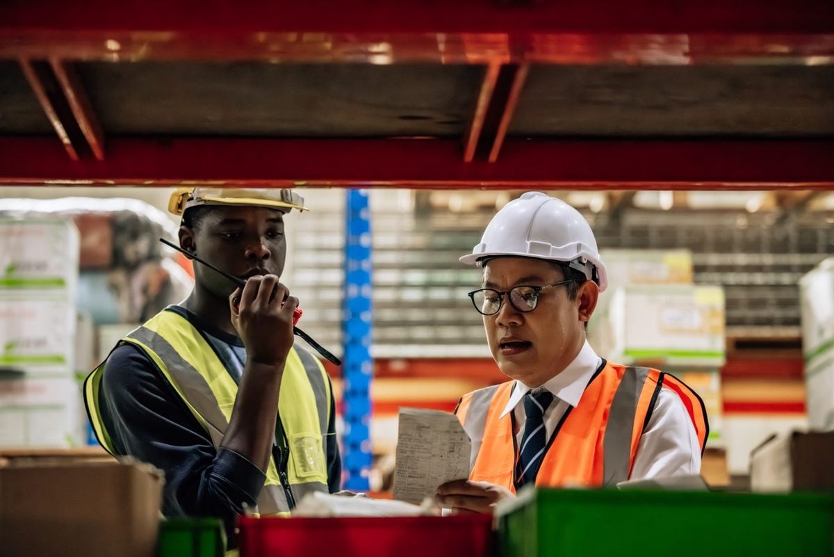 Two warehouse workers checking a printed list together, one in a hard hat reading it, the other speaking into a two-way radio