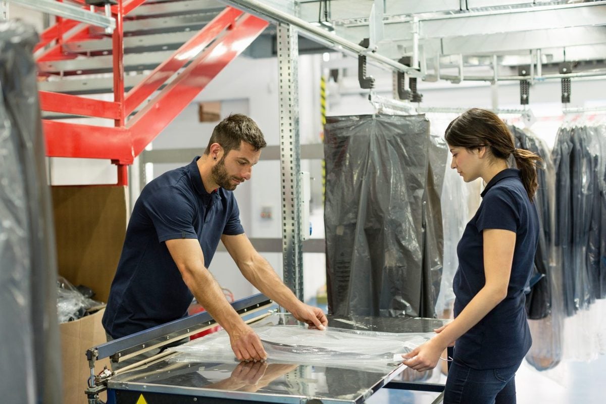 Two workers in navy polo shirts drawing a clear film sheet across a sealing bed on a garment packing line
