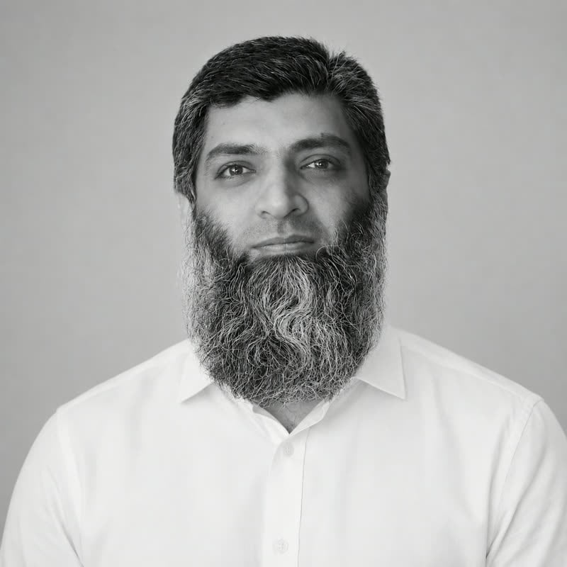 Black and white portrait of Mudassir Iqbal, grey beard, in a white shirt against a plain wall