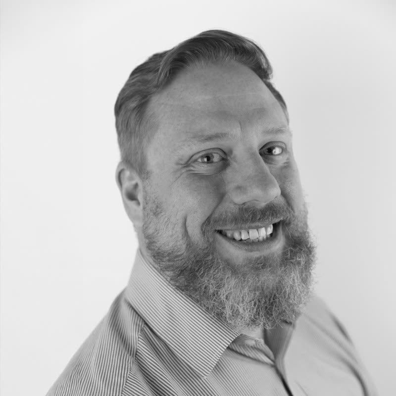 Black and white portrait of Sean Brannon, bearded and smiling, in a fine-striped shirt