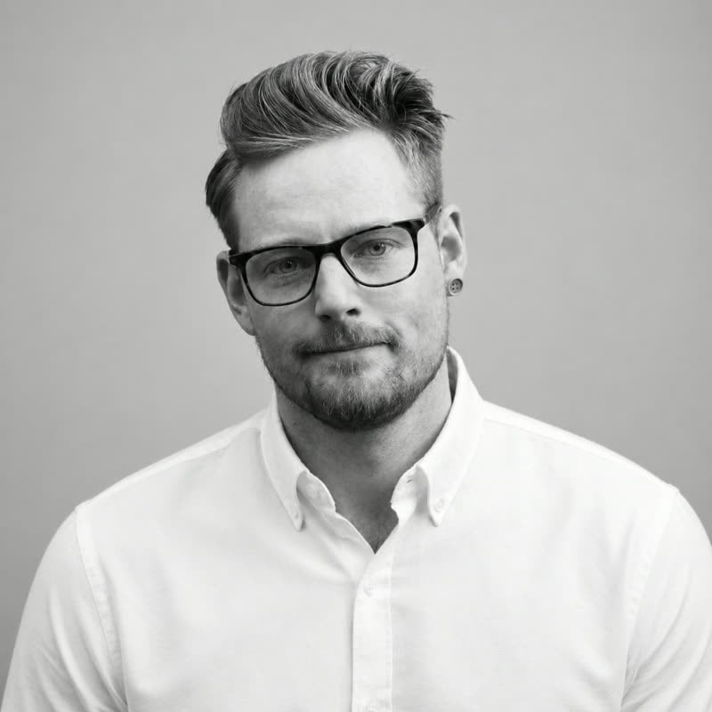 Black and white portrait of Simon Kay in dark-framed glasses and an open white oxford shirt