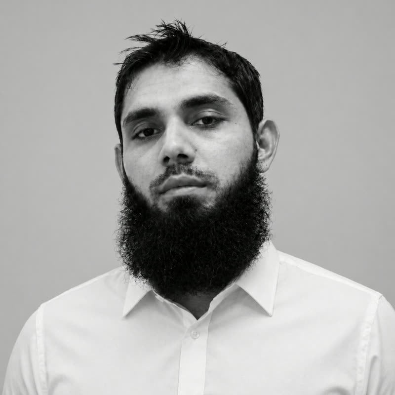 Black and white portrait of Usman Shoukat, full dark beard, in a white shirt against a plain wall
