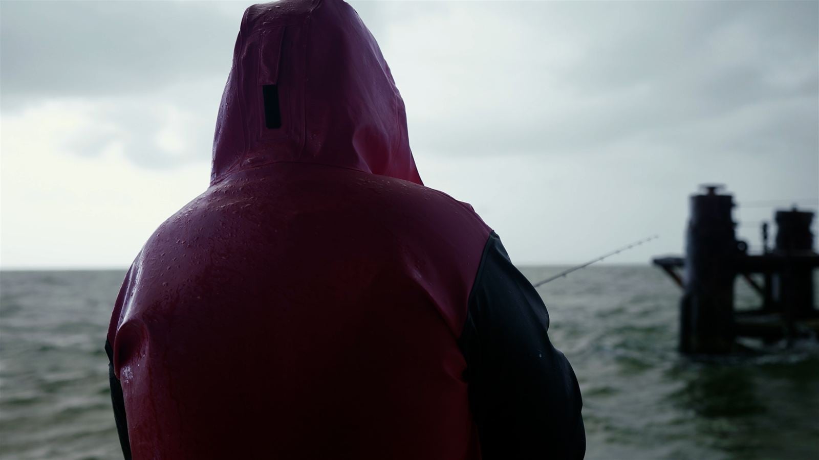 Angler in wet-weather gear fishing from a pier in heavy weather