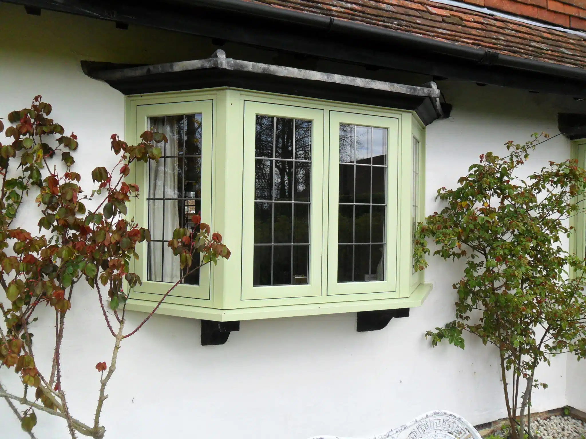 A sage-green bay window installation on a whitewashed cottage with climbing roses