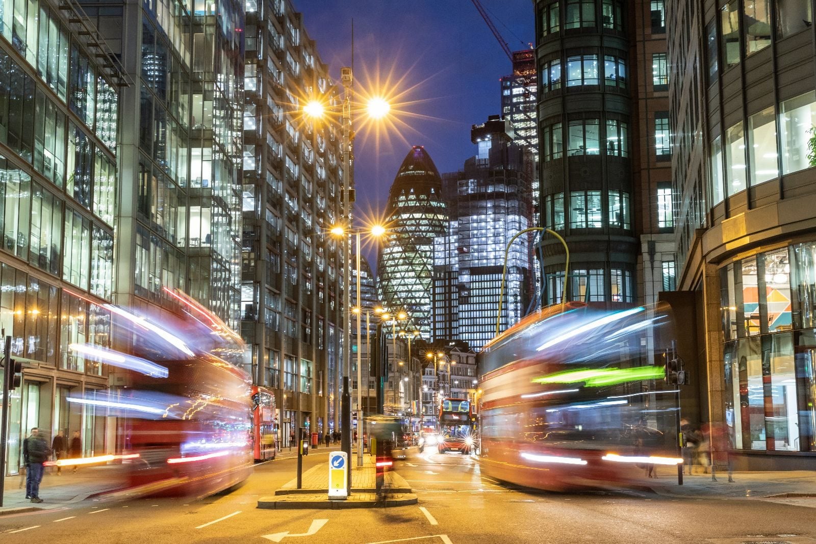 London buses passing in motion blur at dusk, the Gherkin lit up between office towers