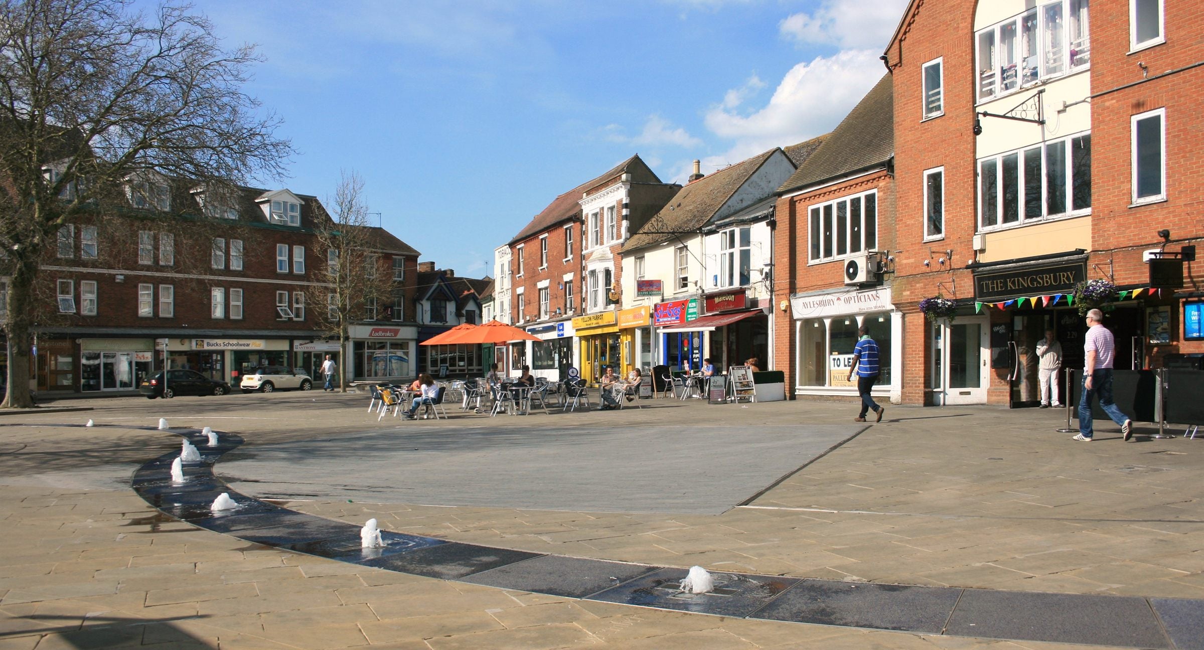 Kingsbury Square in Aylesbury on a bright day, fountains playing in the paved square and cafe parasols outside the shops
