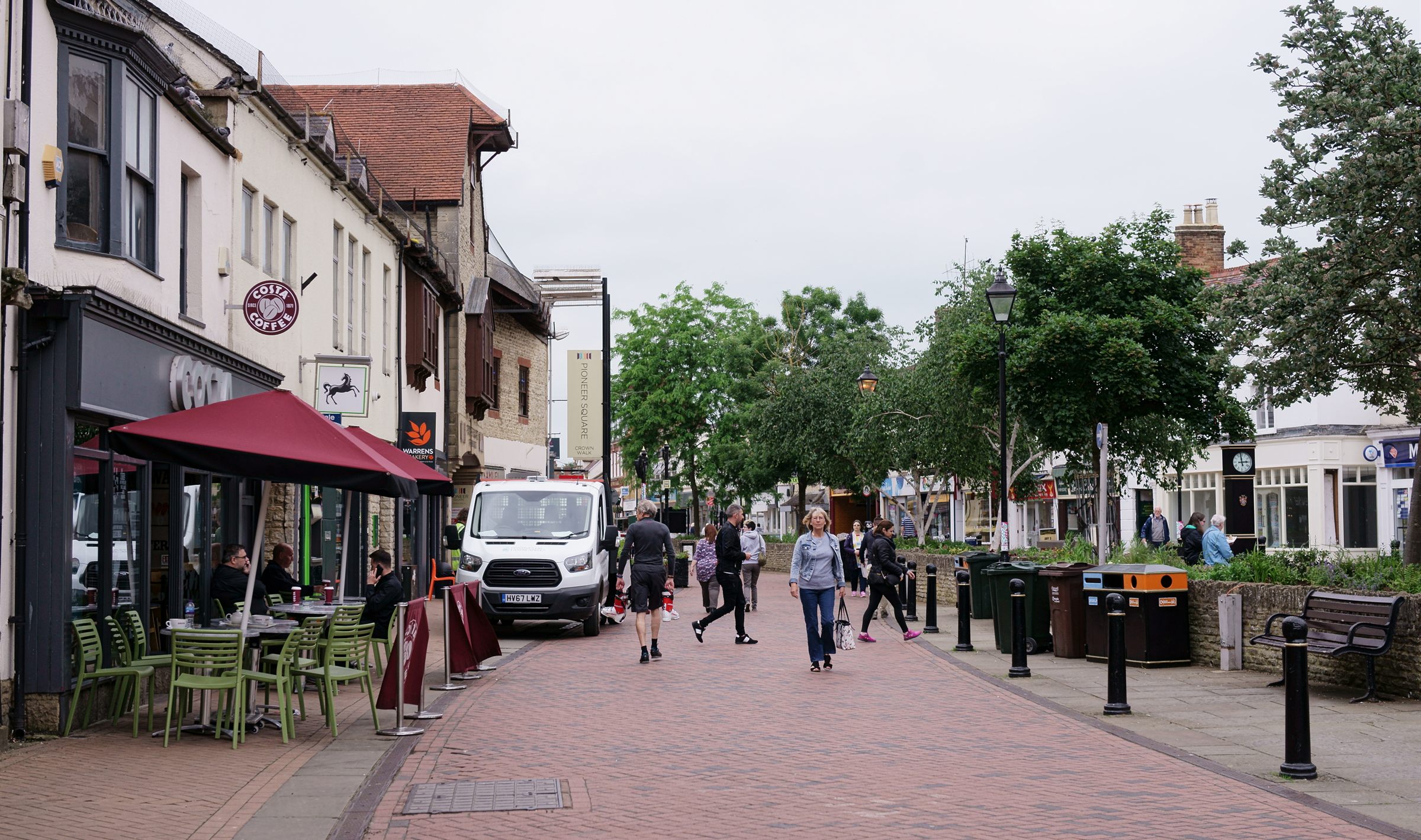 Sheep Street in Bicester, people crossing the paved street between the cafe parasols and the young trees