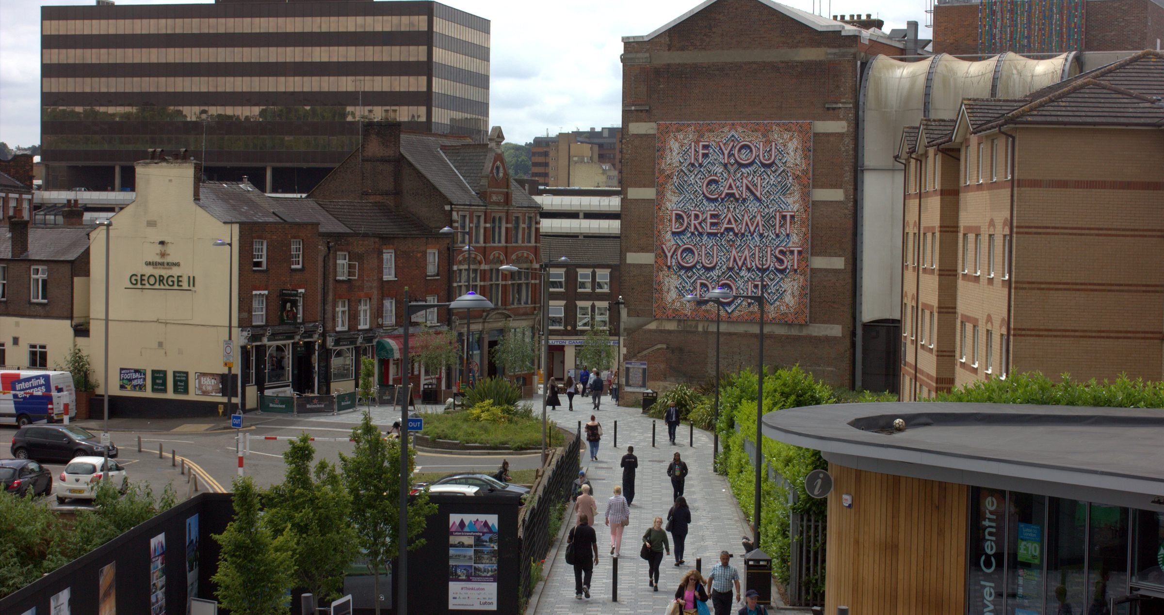 Luton town centre from the station exit, shoppers on the walkway heading towards the mall past a giant painted mural