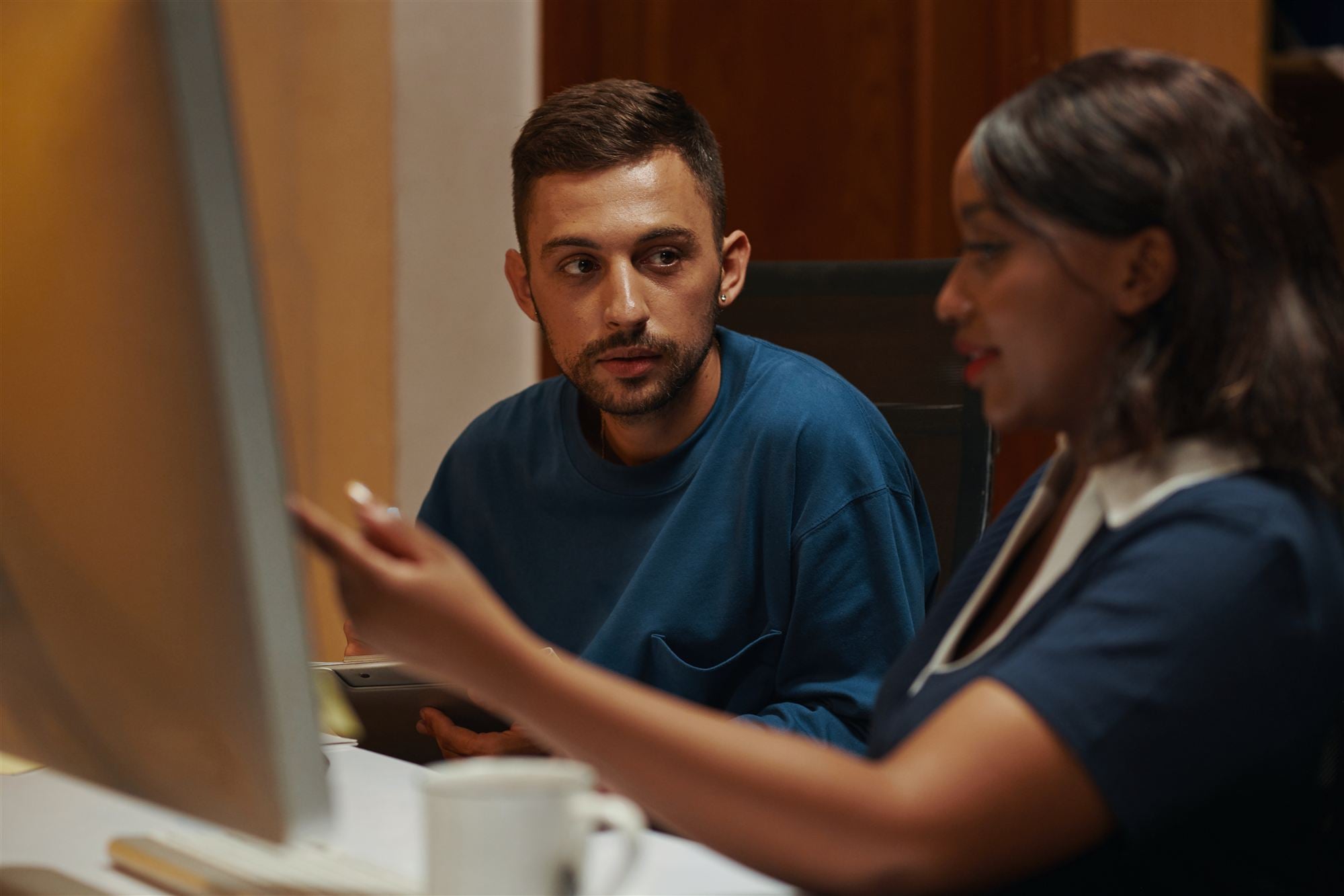 Two people at a studio desk in warm lamplight, one gesturing at a monitor while the other listens, mugs and a tablet on the desk