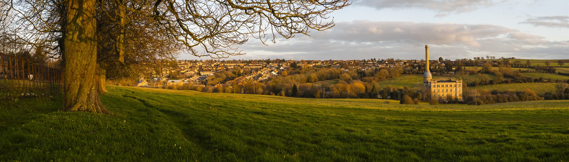 Chipping Norton at golden hour seen across a sloping meadow, the Bliss Tweed Mill chimney standing above the rooftops
