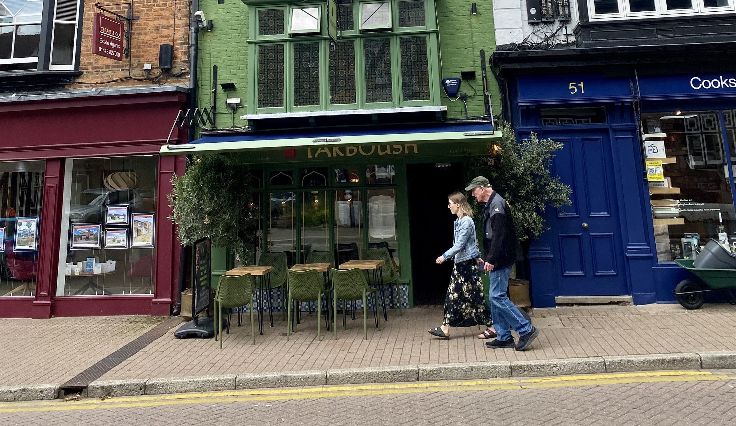 Shopfronts on Tring High Street, a couple walking past the green-fronted Tarboush cafe between the maroon and blue shops either side