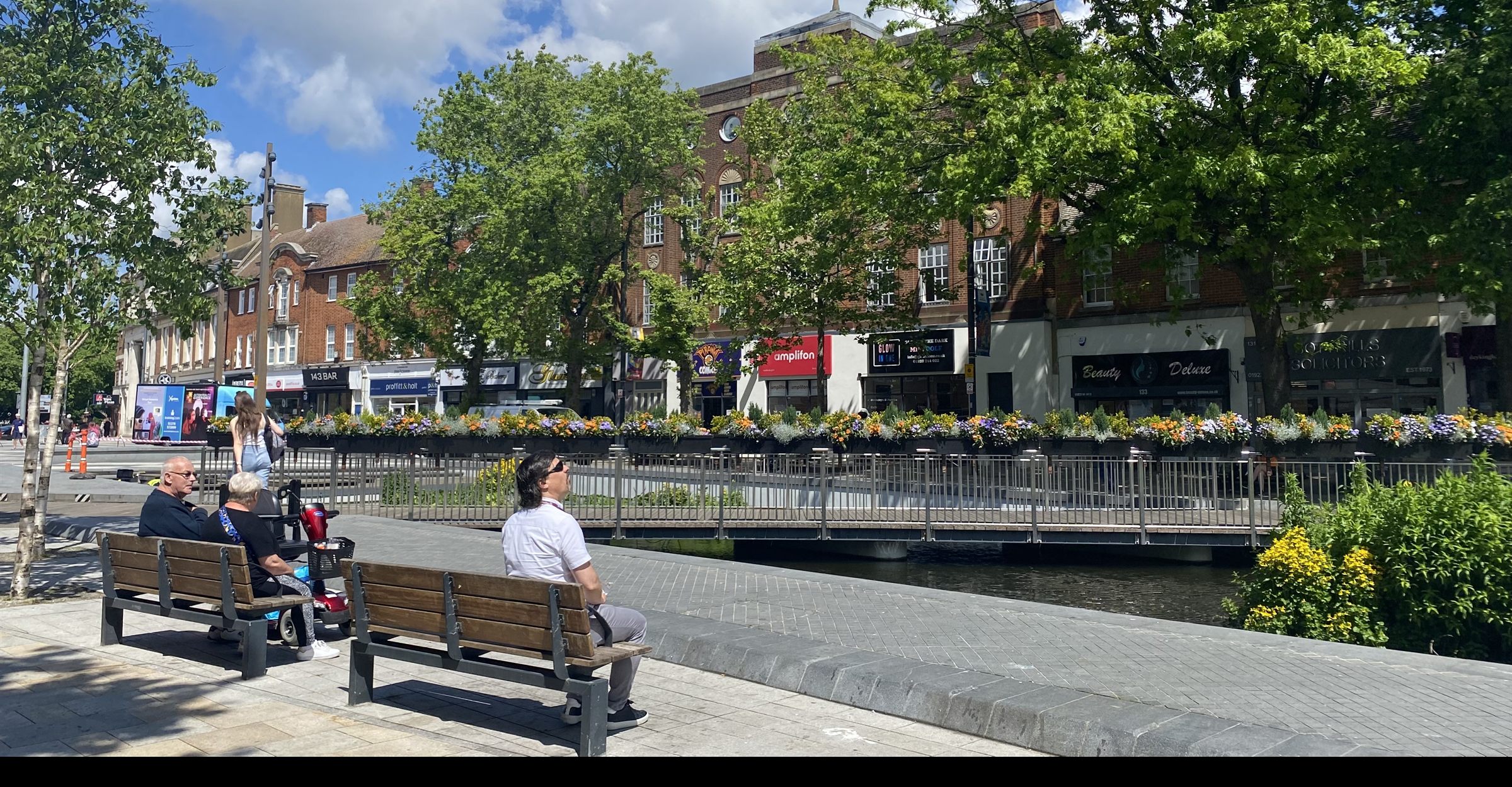 The pond on Watford High Street ringed with flower boxes, people resting on the benches under mature trees