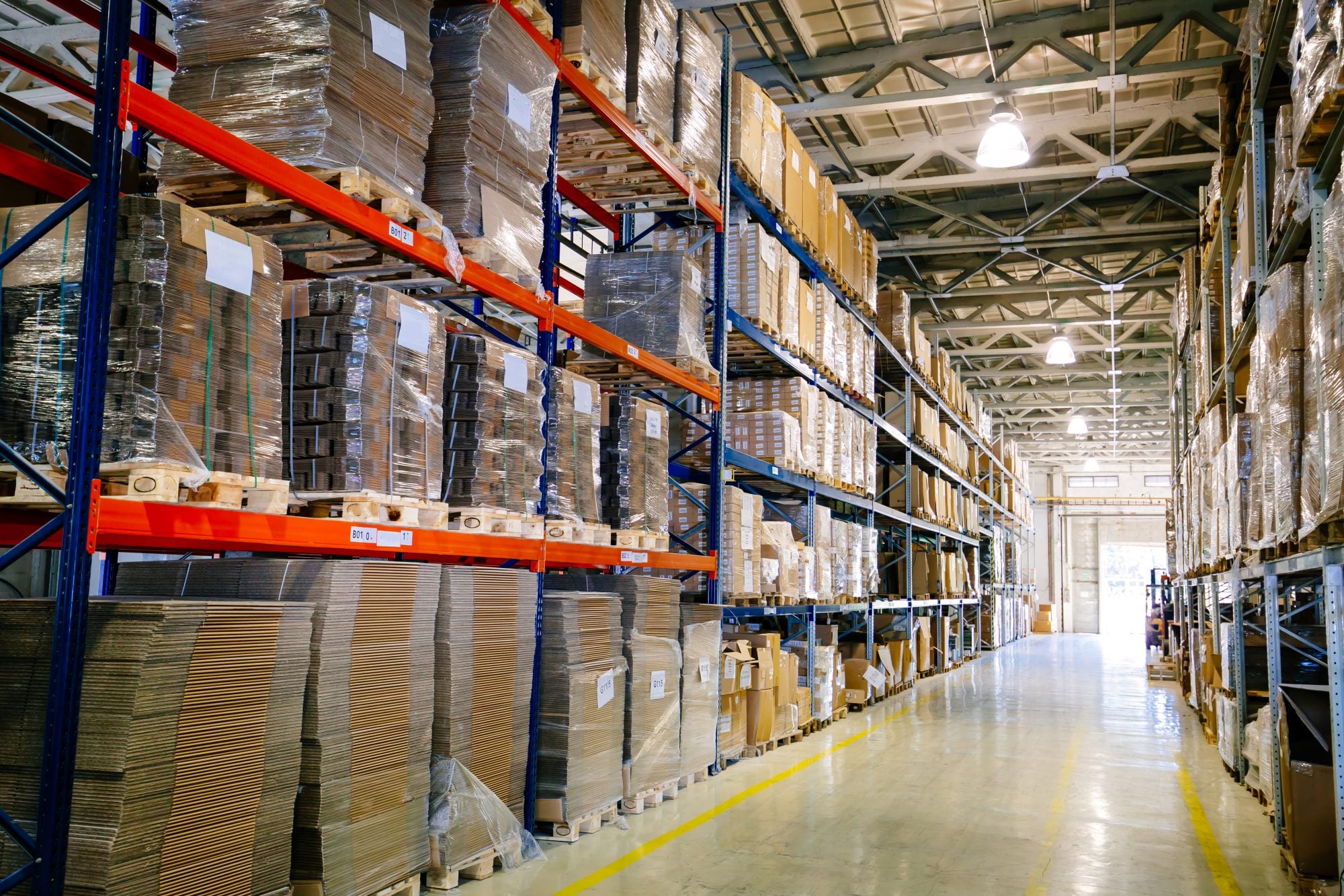 A warehouse aisle of pallet racking stacked to the roof with flat-packed cardboard and shrink-wrapped cartons, a yellow floor line running to the open loading doors at the far end