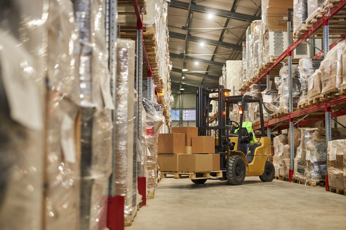 A forklift carrying boxed pallets down an aisle of shrink-wrapped racking