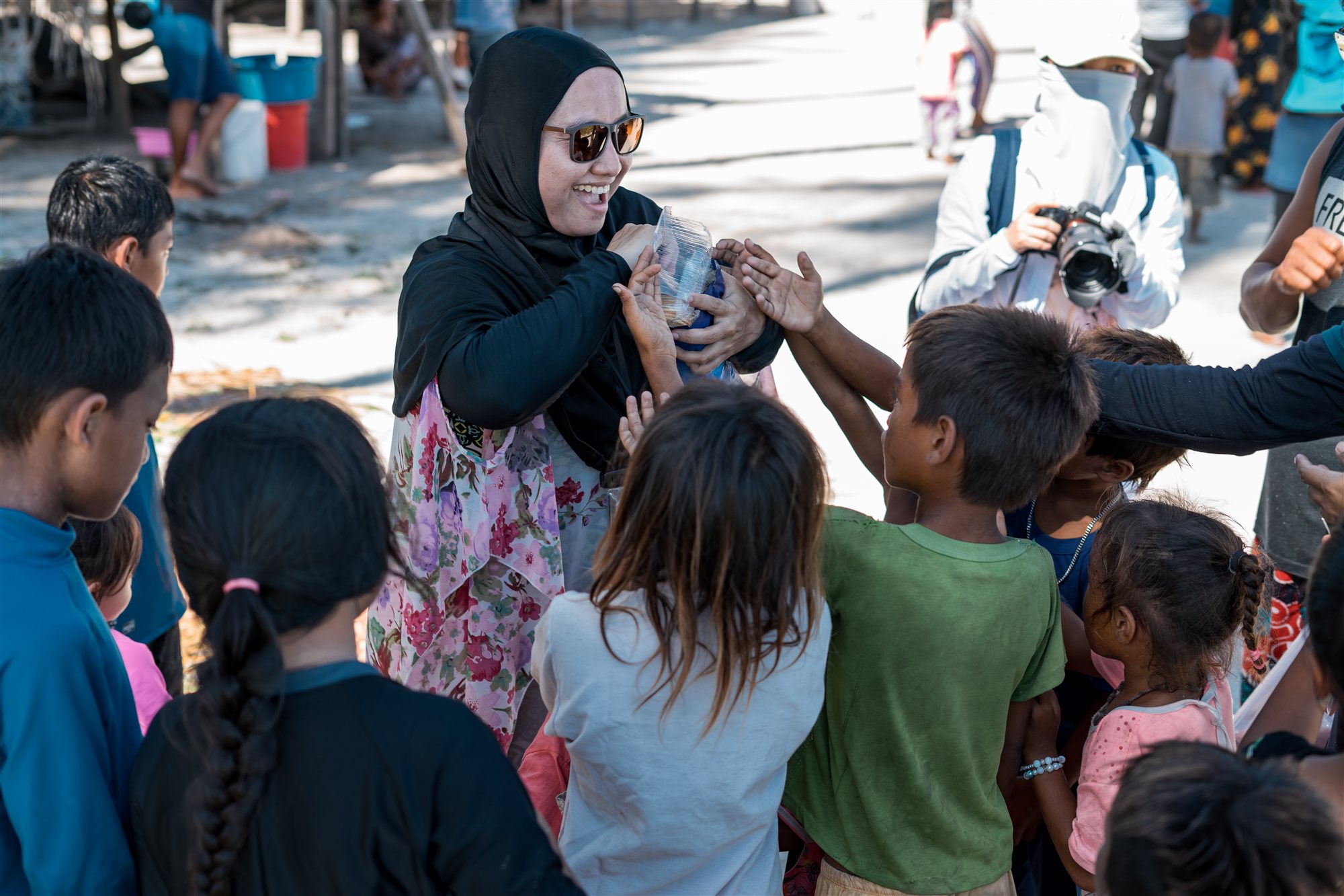 A woman in a hijab handing a wrapped food parcel into a crowd of reaching children on a village street