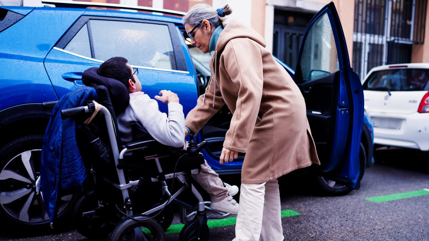 A woman leaning down to help a young man in a wheelchair beside an open car door on a wet street