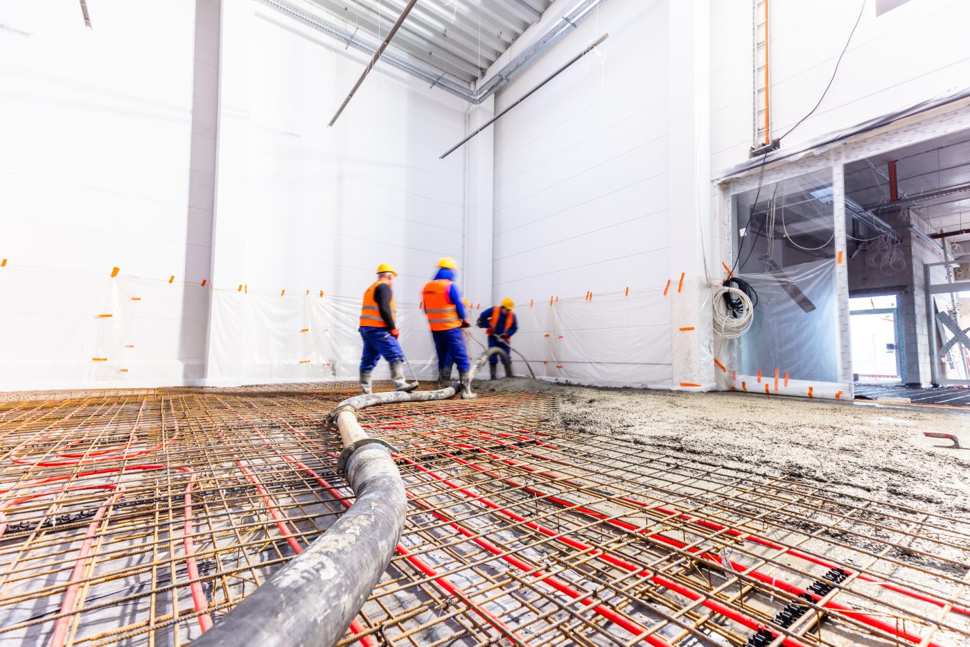 Three workers in orange vests pumping screed over rebar mesh and underfloor pipework