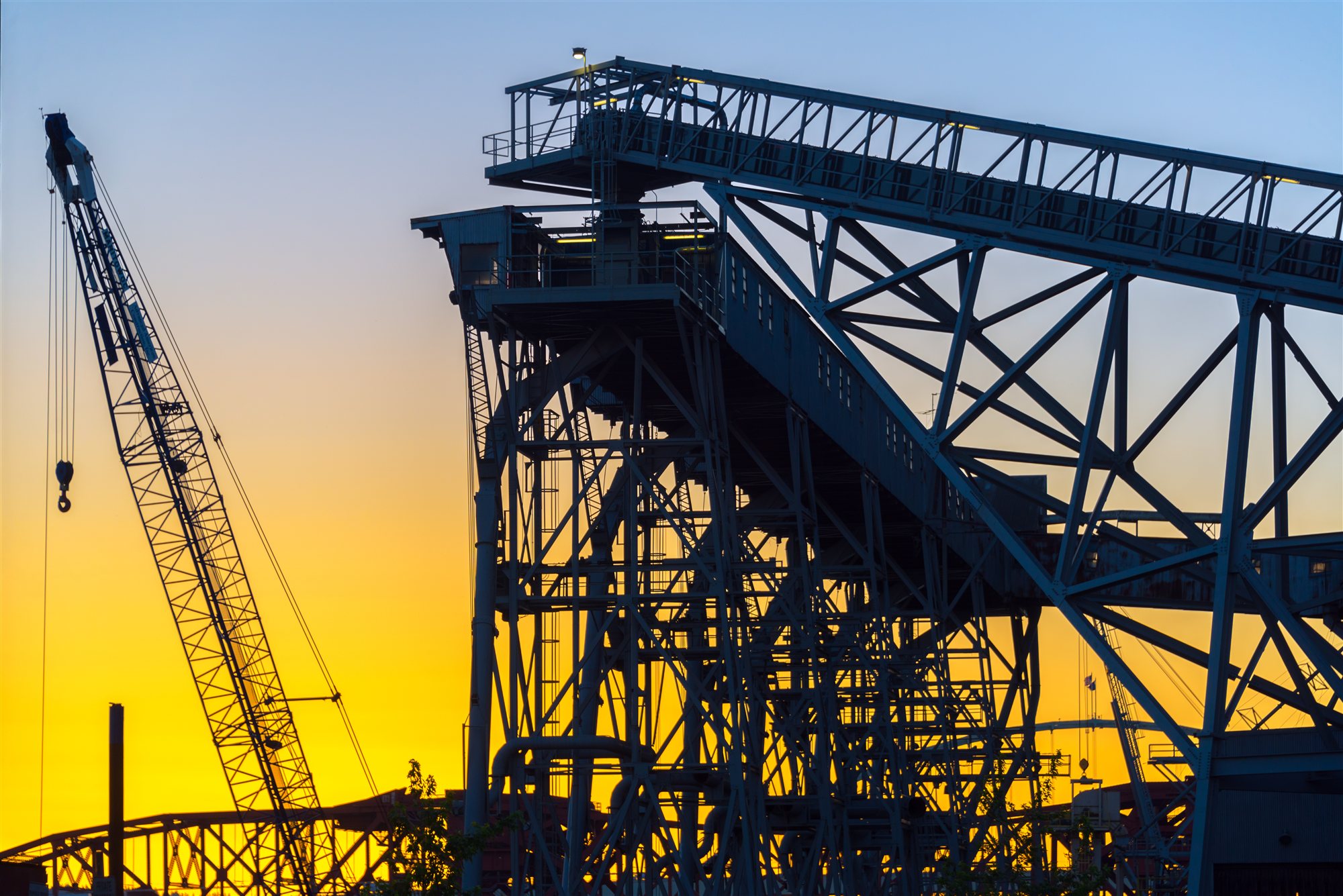 A silhouetted lattice crane and conveyor gantry against a vivid orange sunset sky