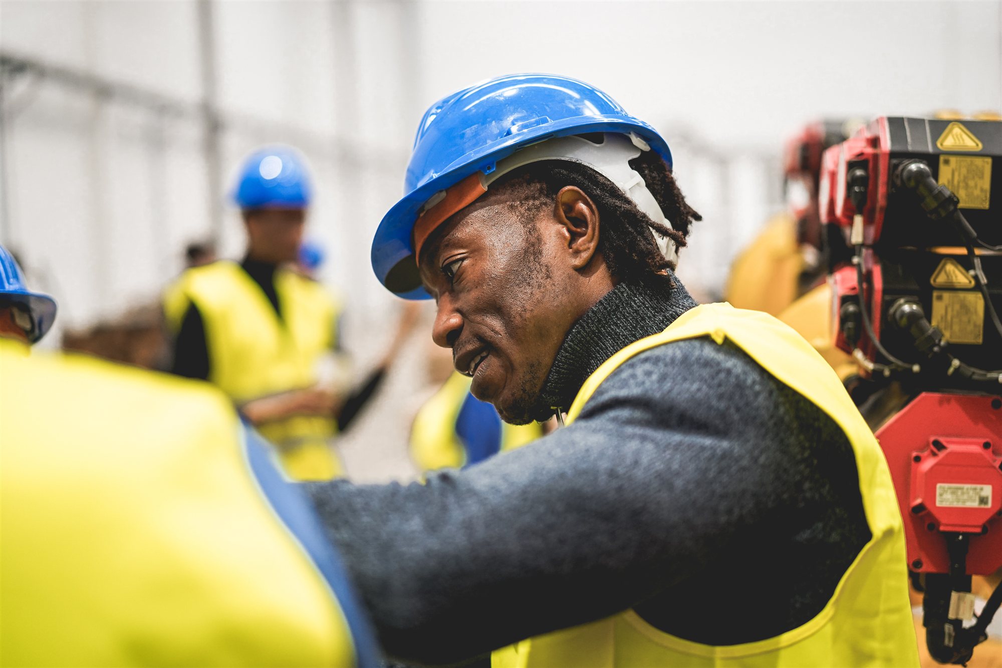 A worker in a blue hard hat and yellow hi-vis leaning over machinery, colleagues blurred behind him