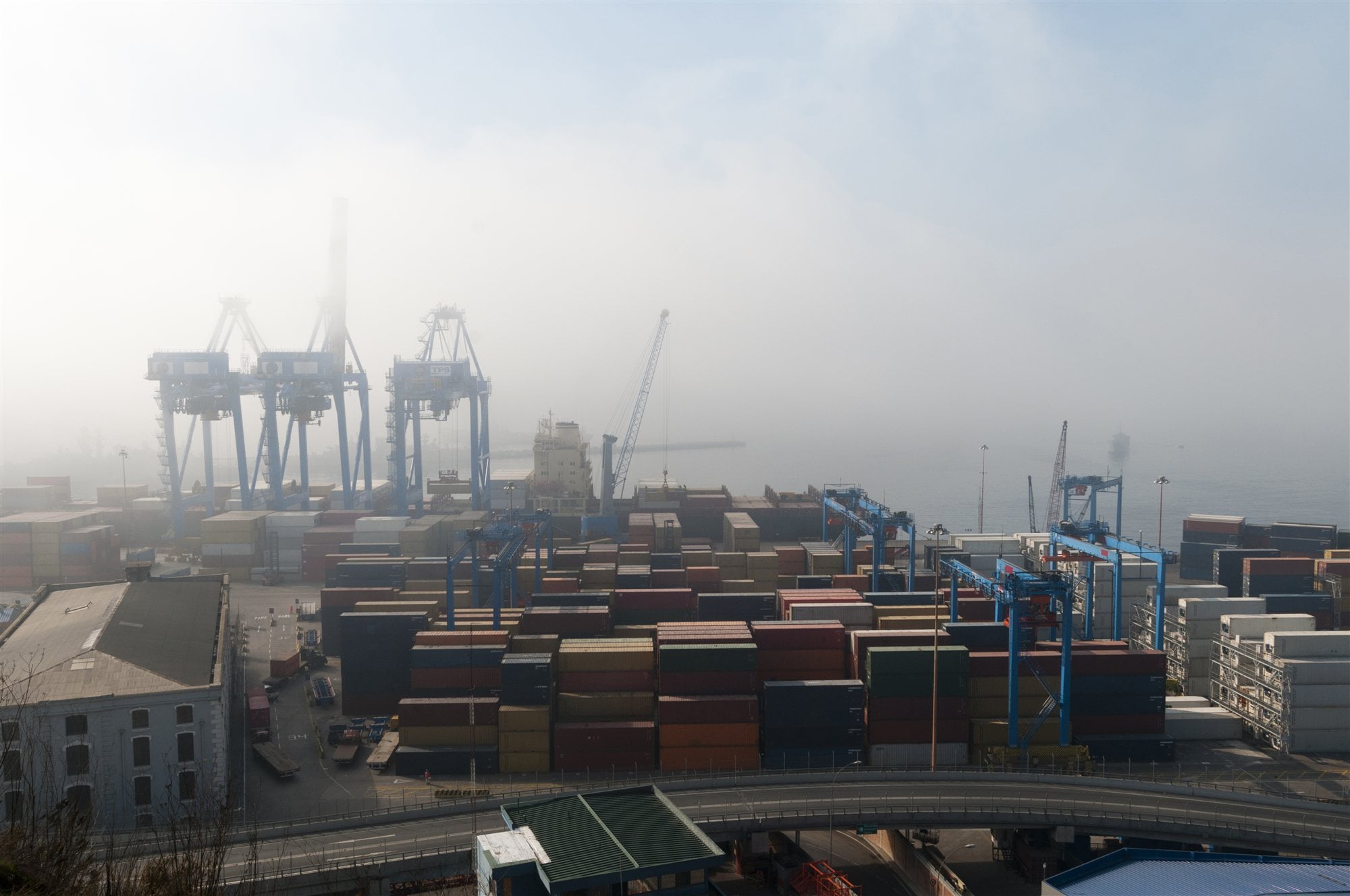 A container port at dawn, blue gantry cranes and stacked containers half lost in sea fog