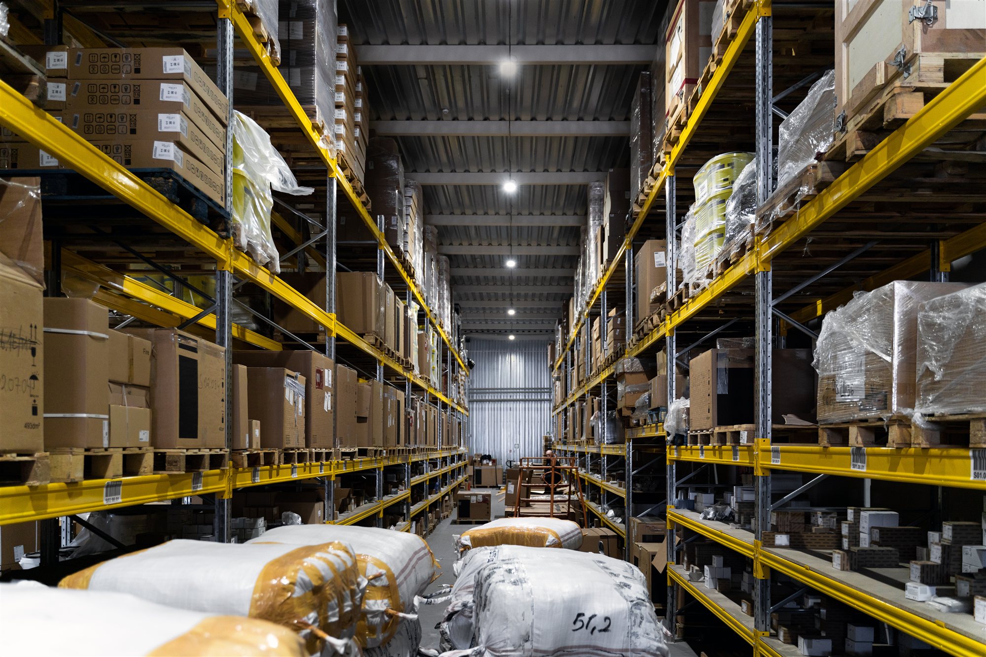 A symmetrical aisle down tall yellow pallet racking loaded with boxes, a single figure at the far end