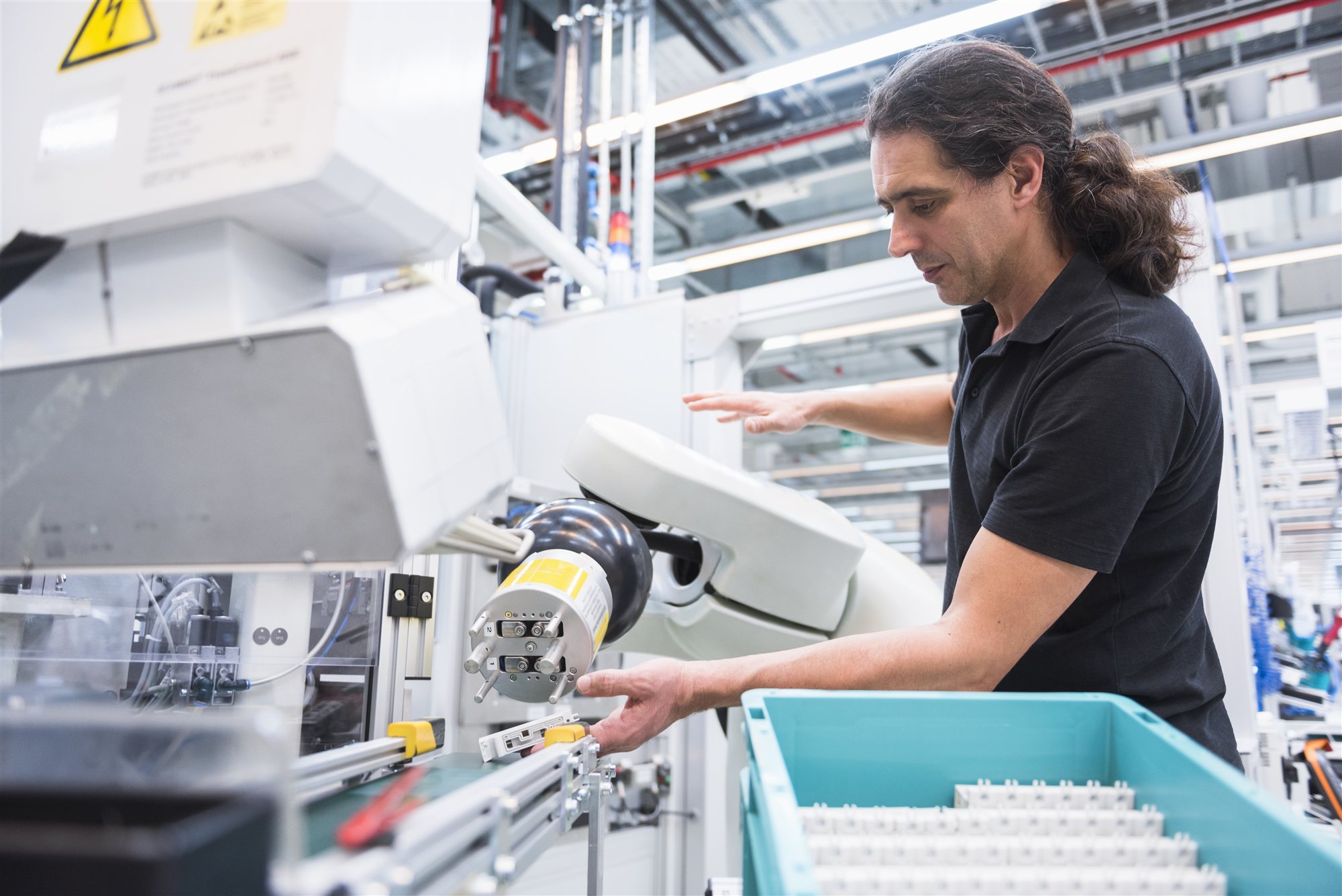 A man reaching into a white robotic pick-and-place cell, feeding small components into a teal crate