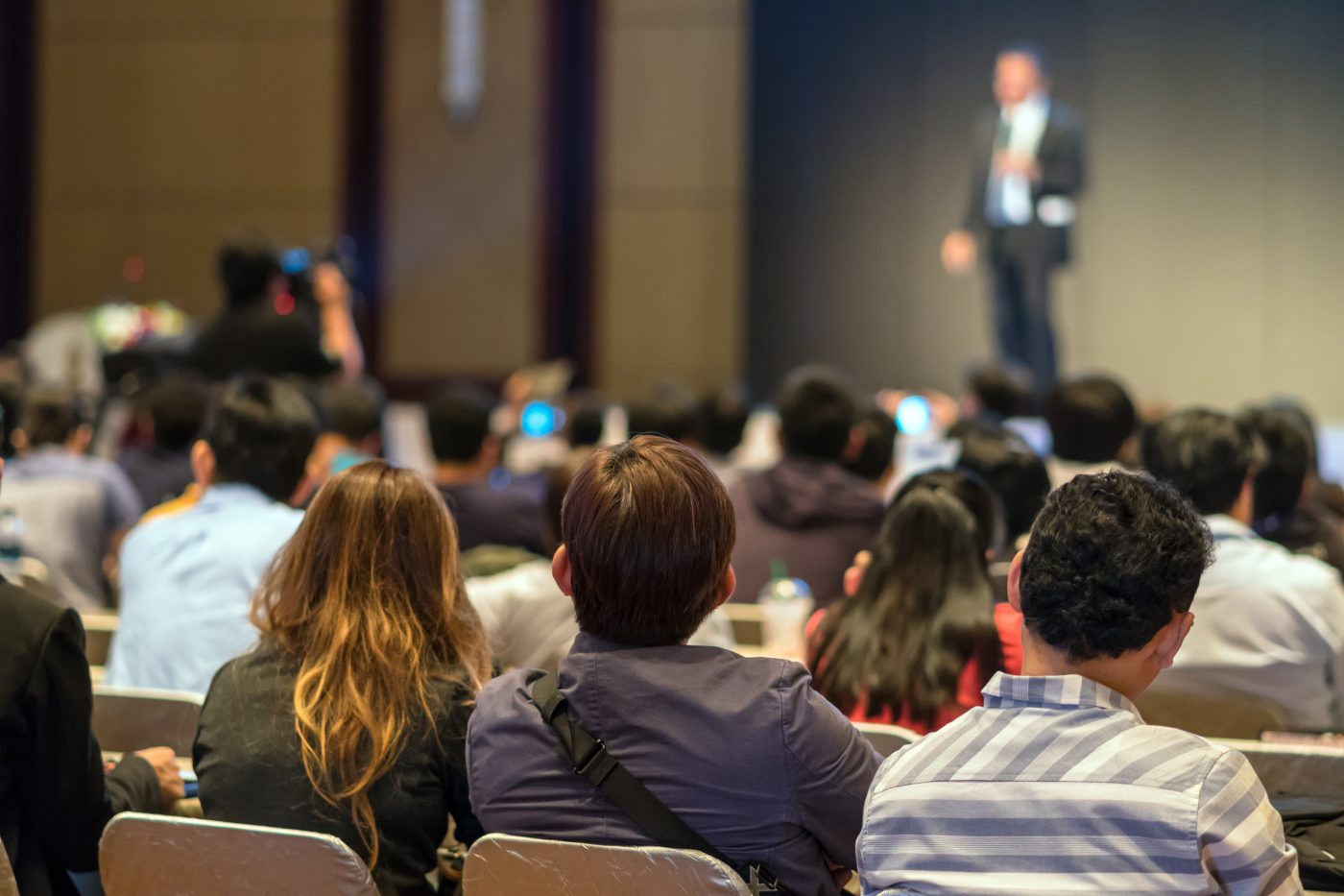 A seated conference audience seen from behind, phones raised toward a lit stage