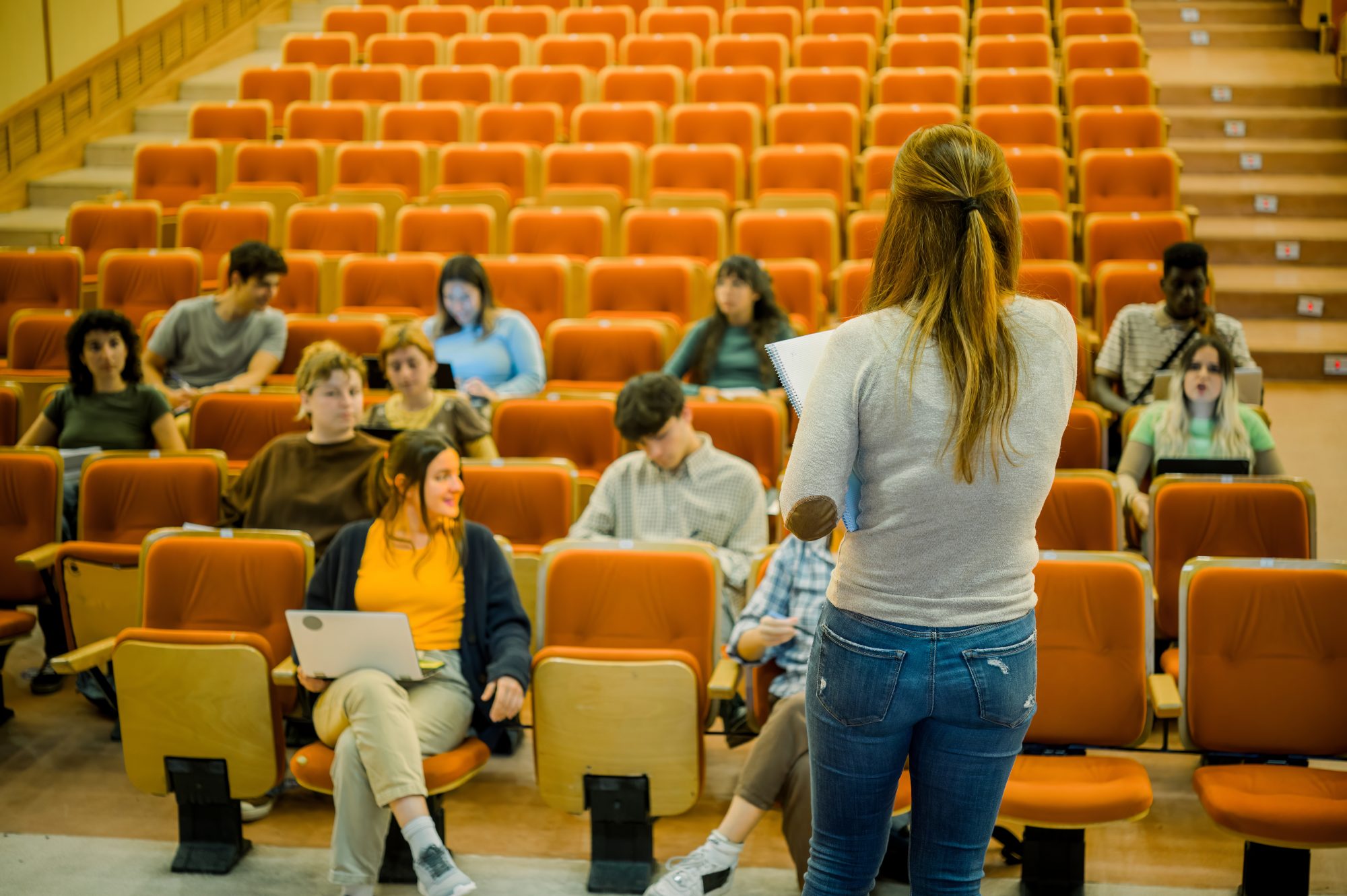 A lecturer with her back to camera addressing students scattered across orange lecture-theatre seats