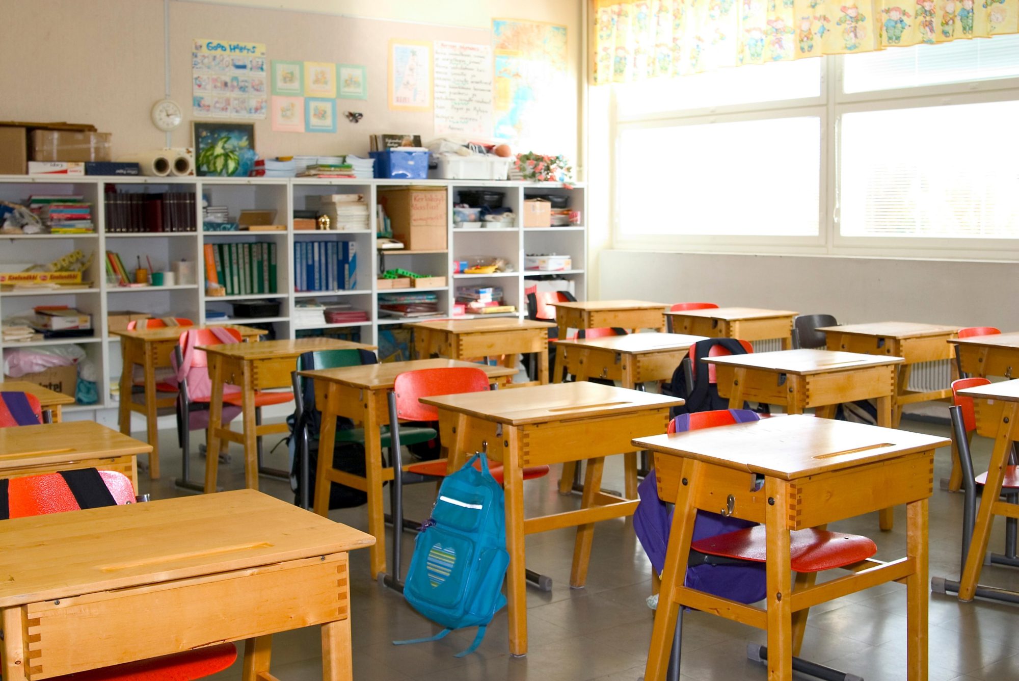 An empty primary classroom of pine desks and red chairs, a blue backpack left on the floor