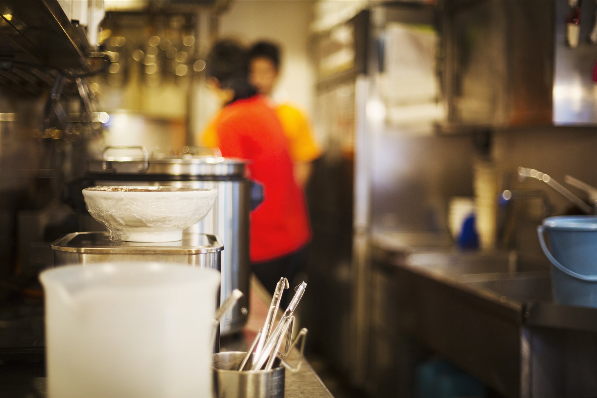 The foreground of a commercial kitchen pass, stock pots and a cling-filmed bowl, two staff working behind under warm light
