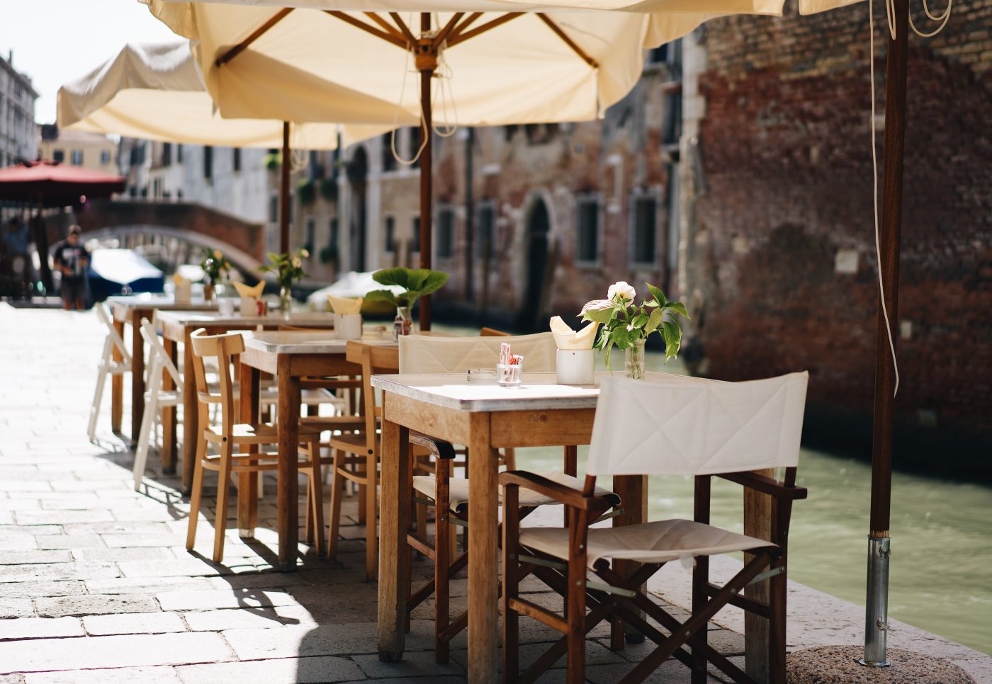 A row of wooden cafe tables with parasols along a canal edge, laid and waiting