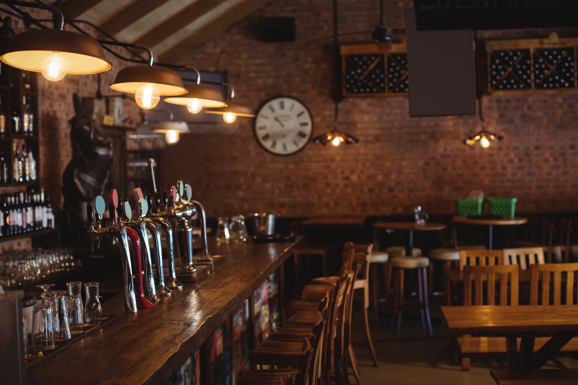 An empty country pub interior, a long dark wood bar with beer pumps under pendant lamps, laid tables to the right