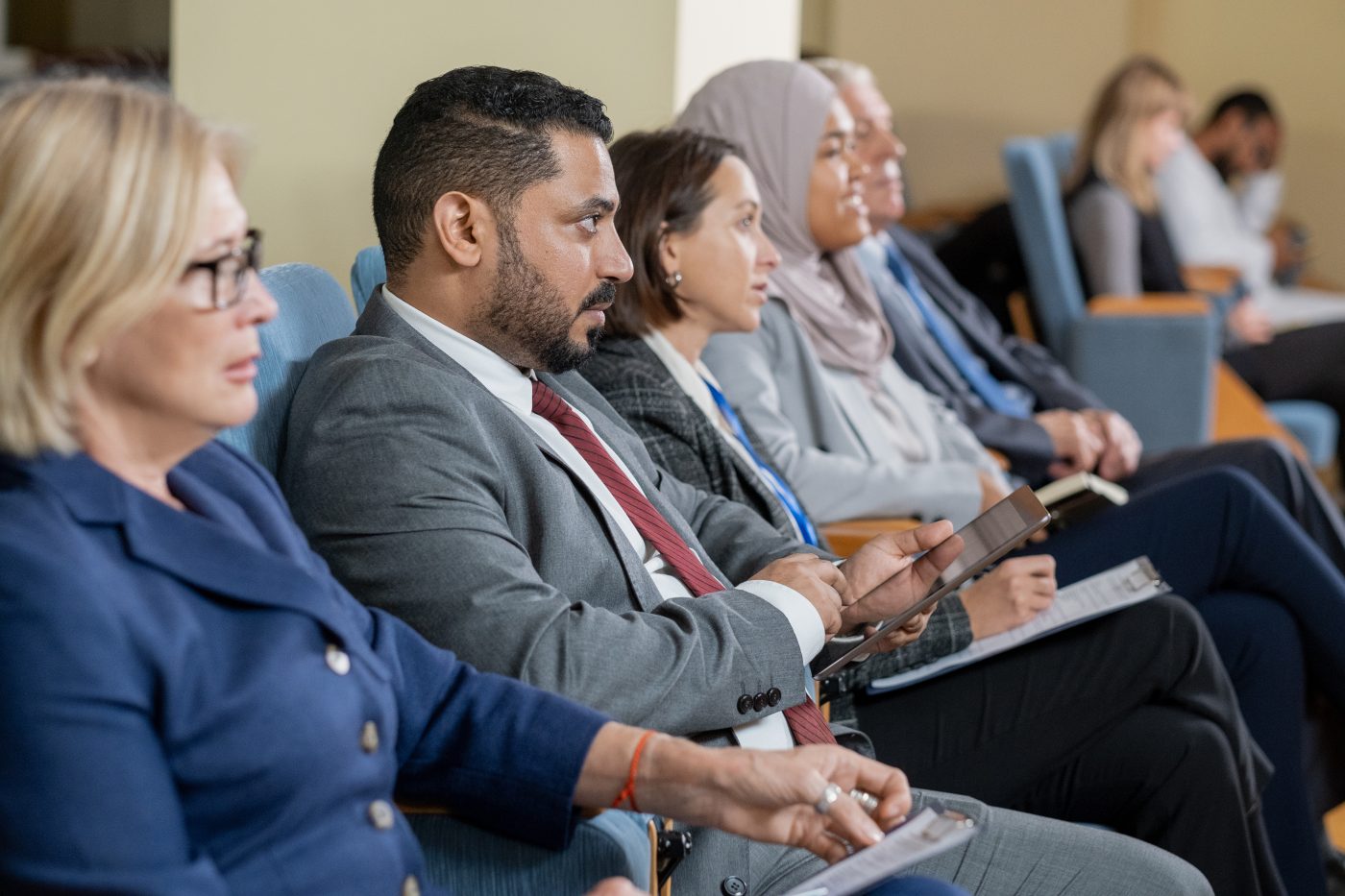 A row of seated delegates in suits listening in a conference room, one holding a tablet