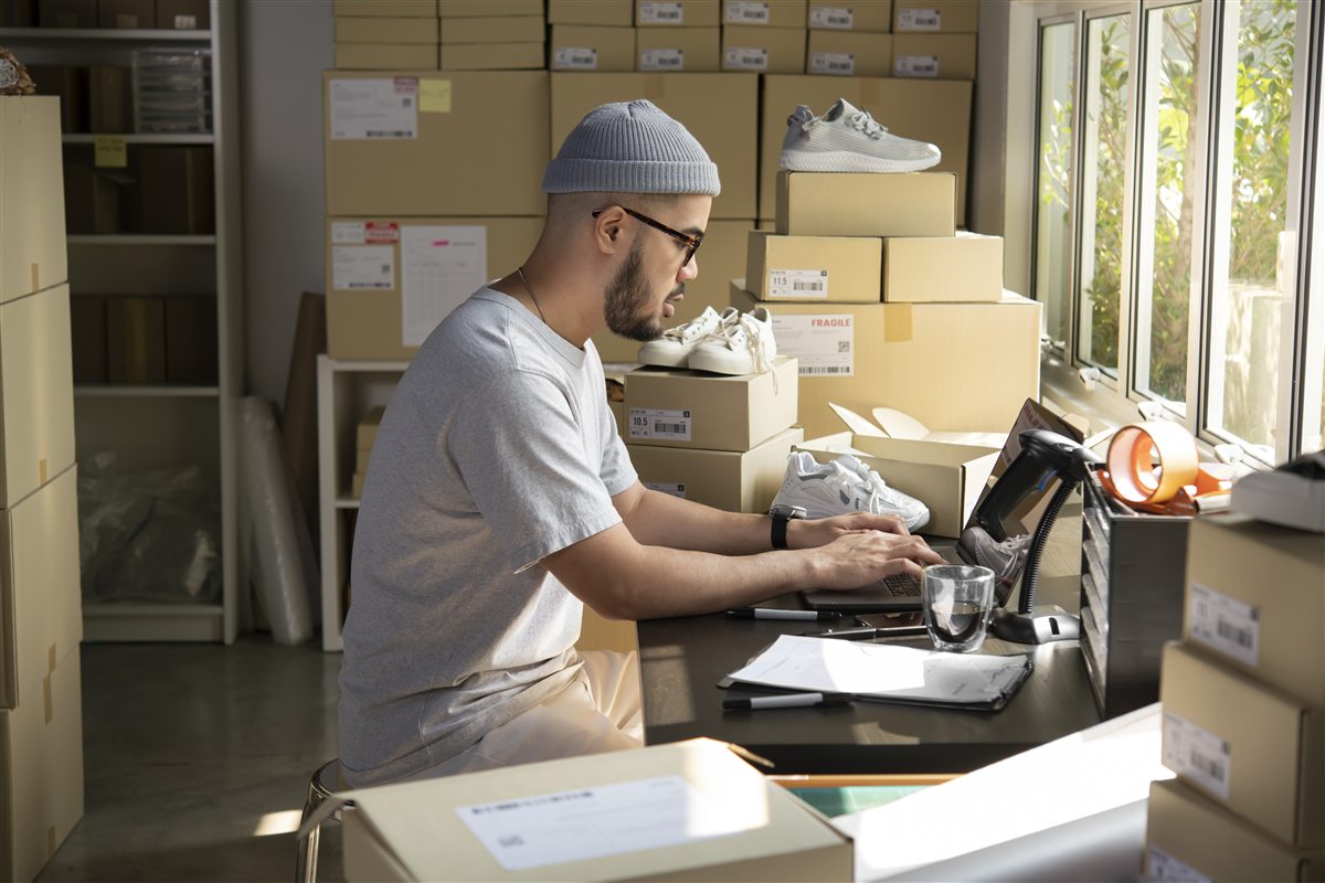 A man in a beanie working at a laptop in a stockroom, shipping boxes and trainers stacked around him