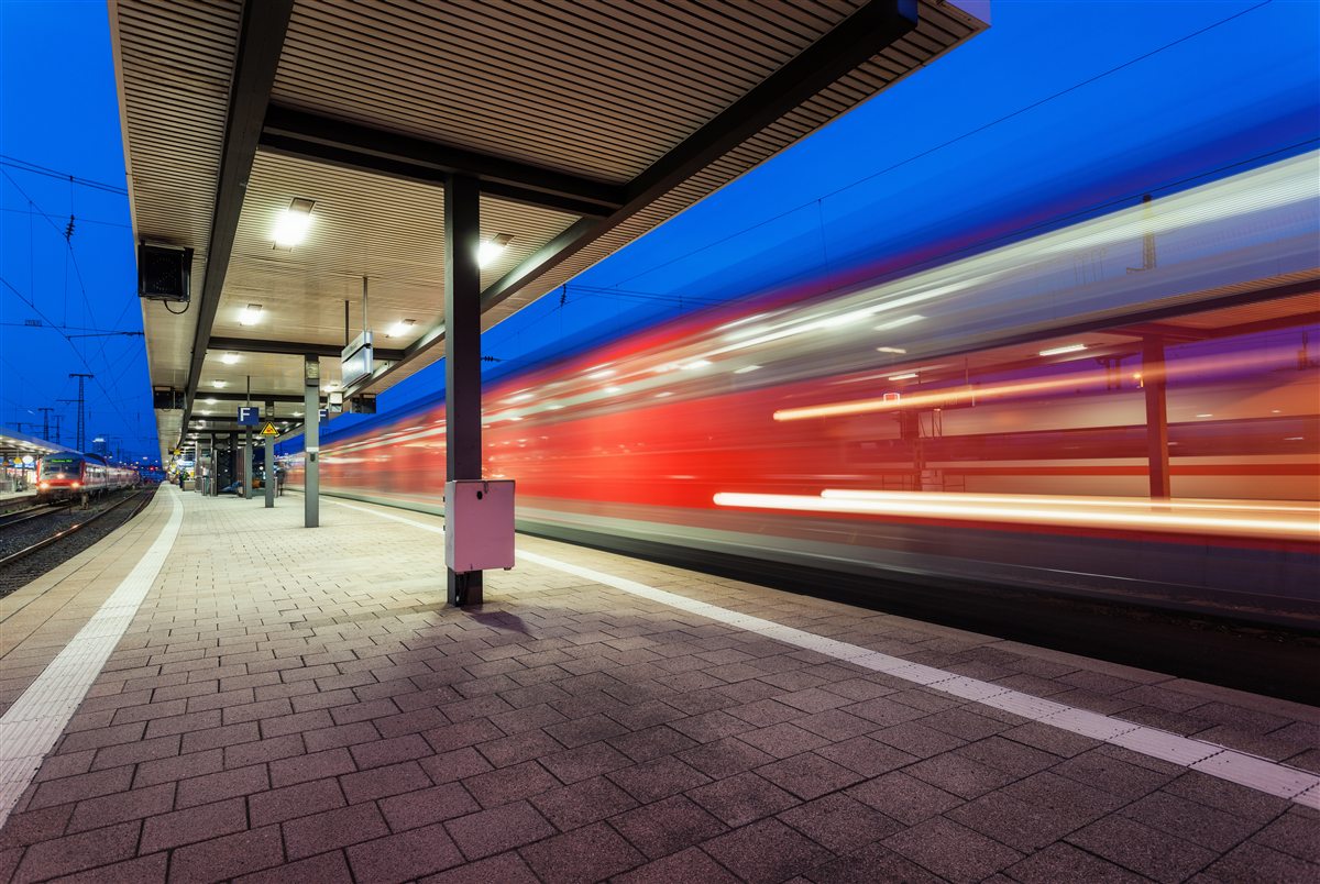 A train streaking past a covered station platform at blue hour, rendered as red and white motion blur