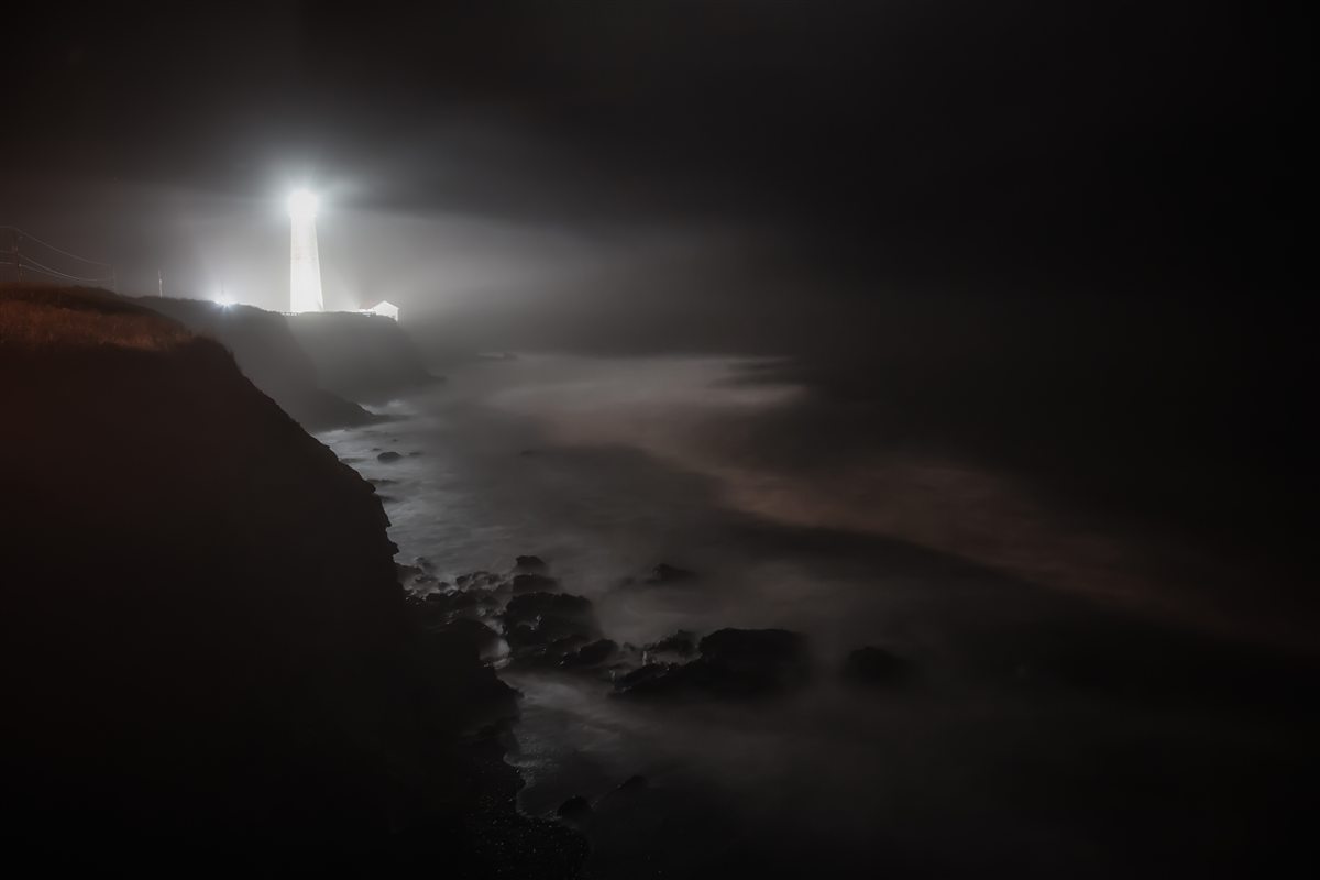 A lighthouse beaming through heavy fog at night, long-exposure surf over the rocks below