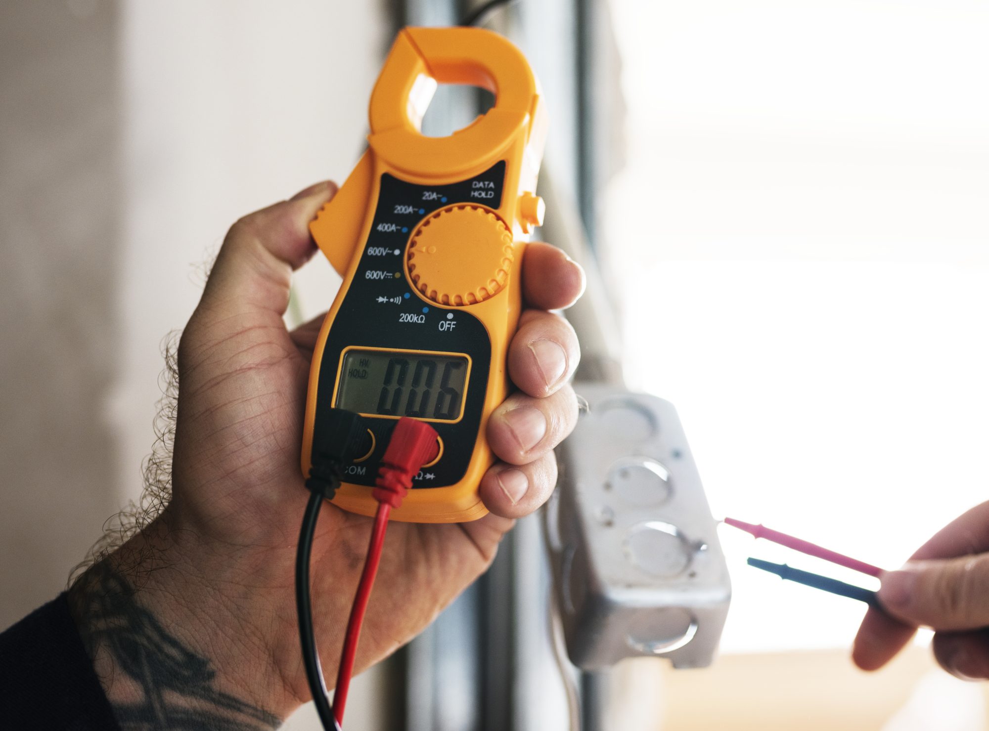 A tattooed hand holding an orange clamp multimeter while the other holds probes to a metal back box