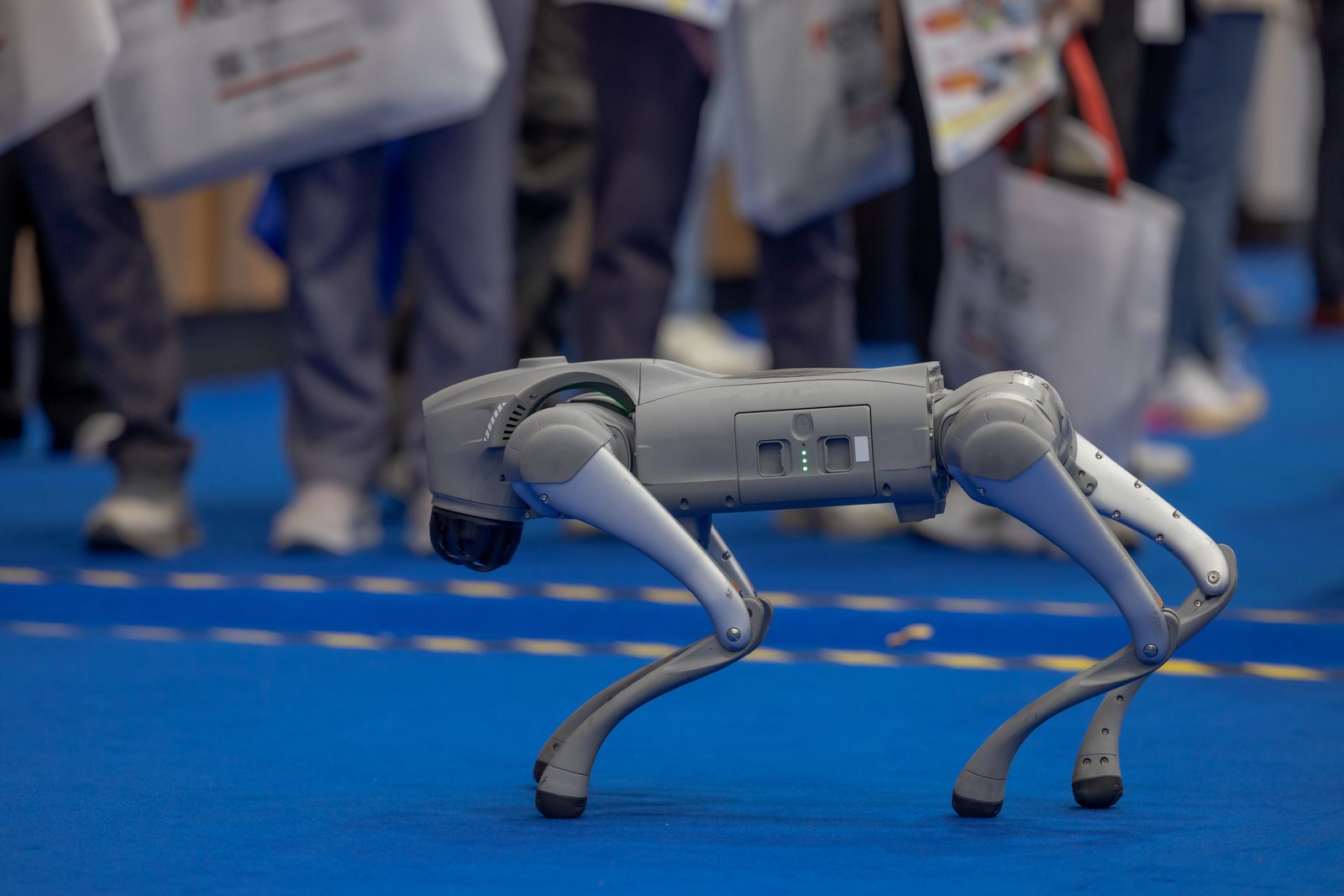 A grey quadrupedal robot walking a blue exhibition carpet, trade-show visitors blurred behind