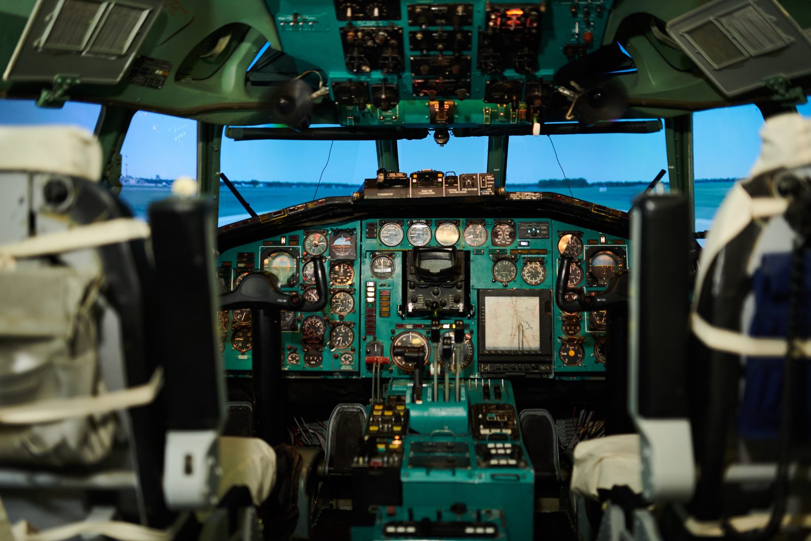 An aircraft cockpit with twin control yokes and banks of teal instrument panels, the runway ahead