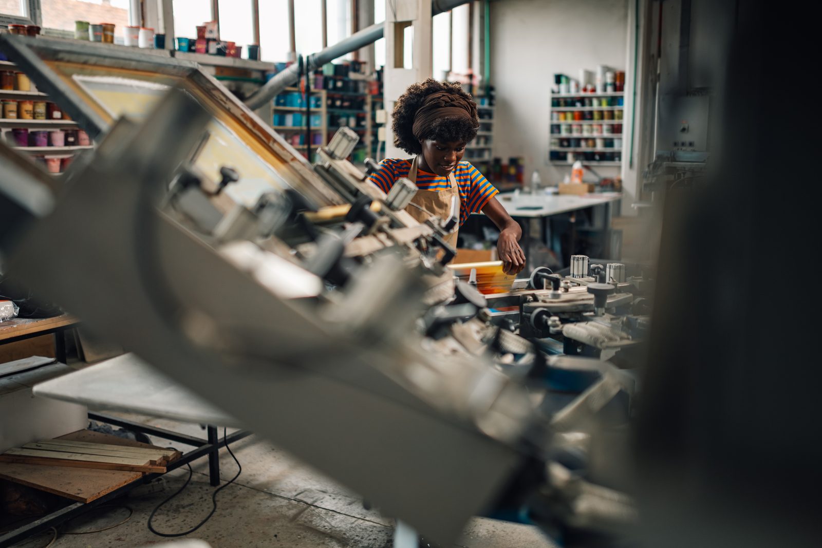 A screen printer in an apron working a silk-screen carousel, shelves of ink pots behind her