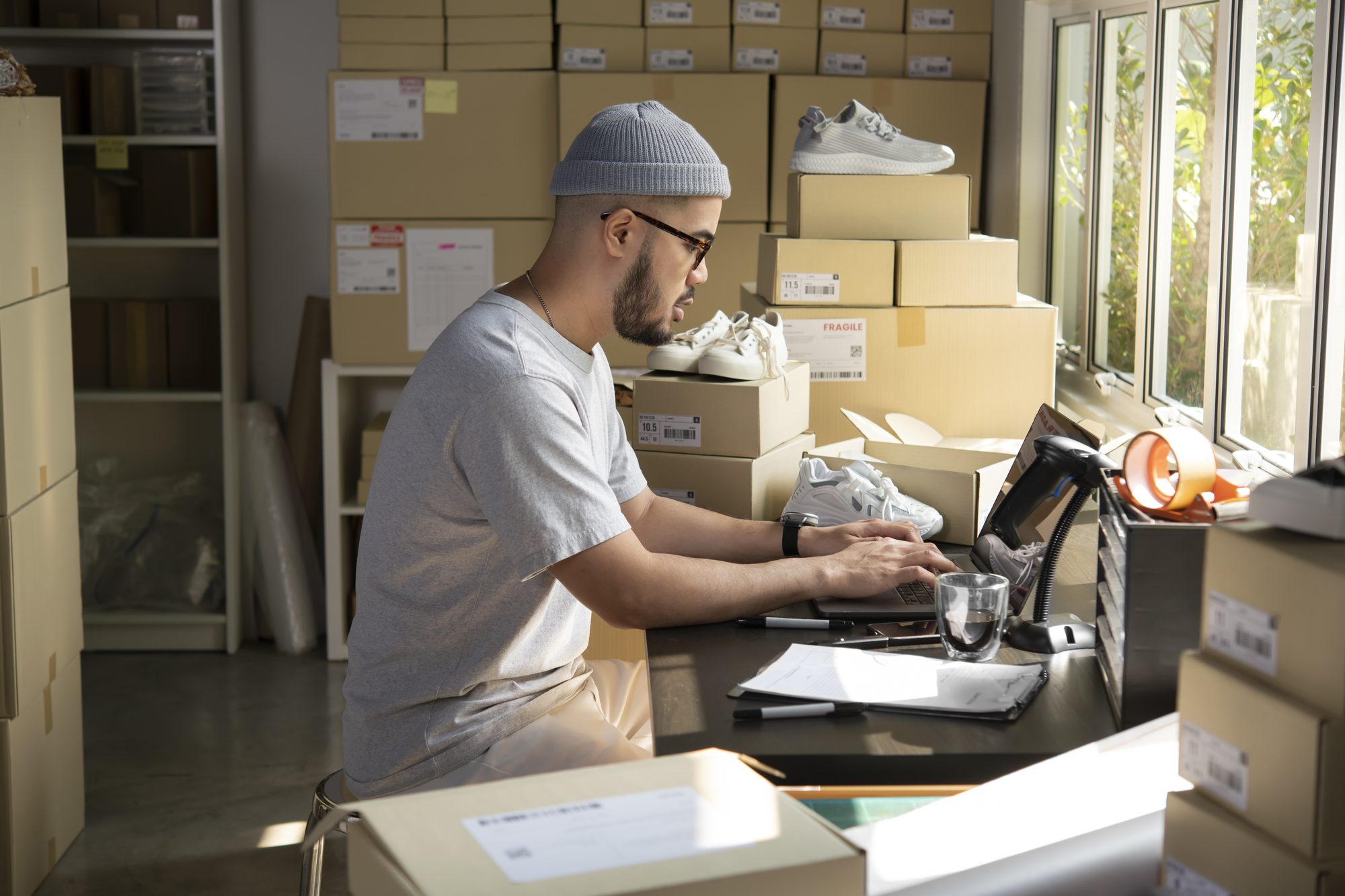 A seller in a beanie updating orders on a laptop, surrounded by stacked shoe boxes and boxed trainers