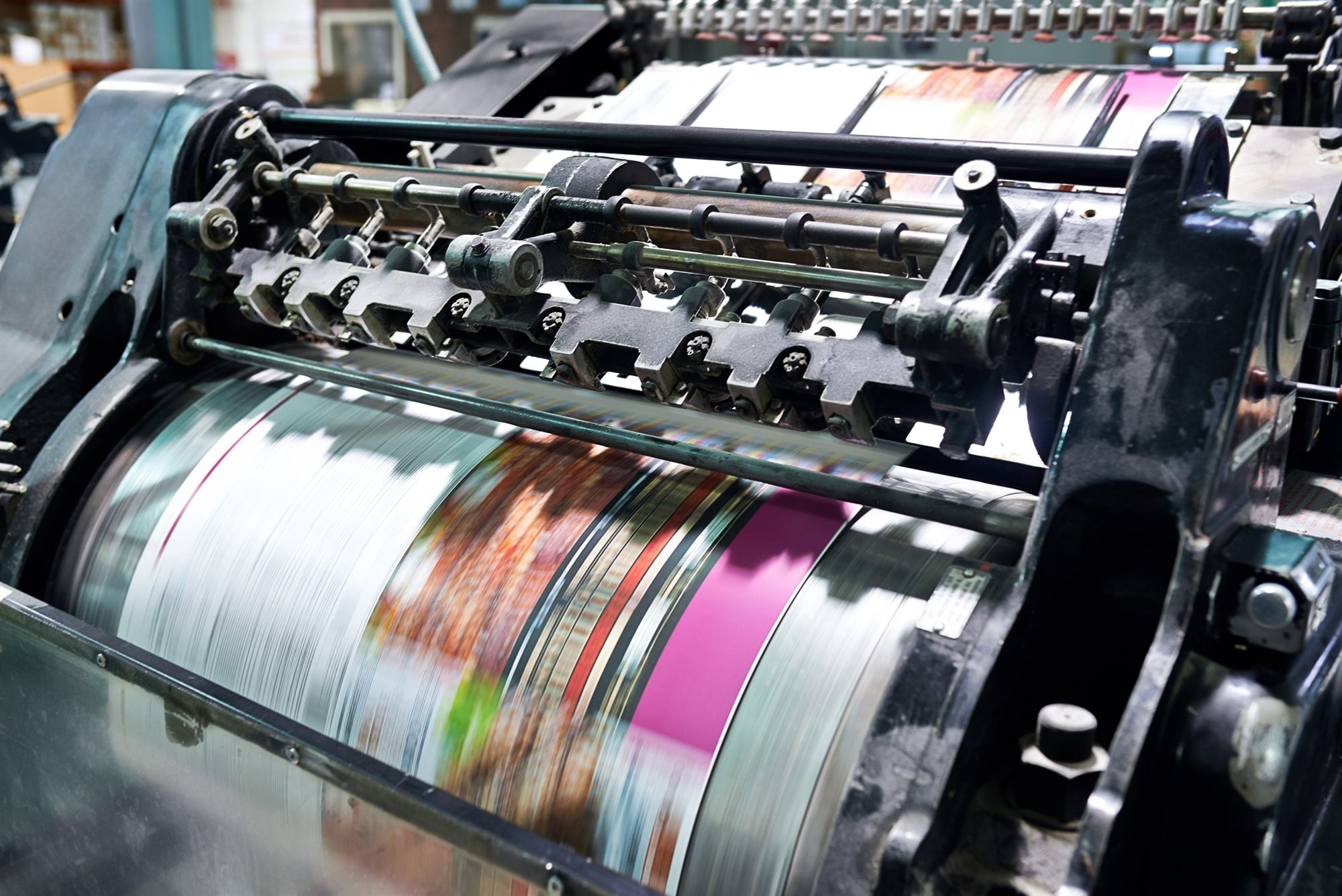 A running offset printing press throwing motion-blurred magenta and cyan bands under the ink rollers