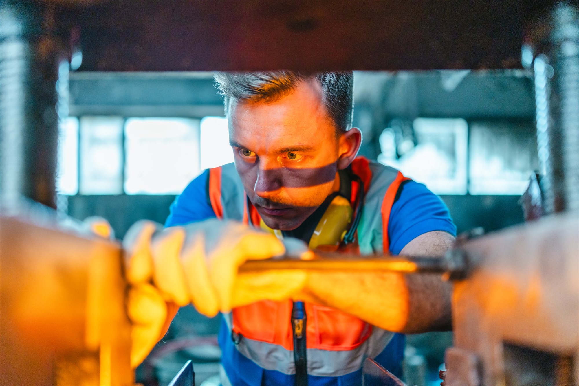 A technician in a hi-vis vest steadying a metal bar in factory machinery, his face lit by a warm glow from below