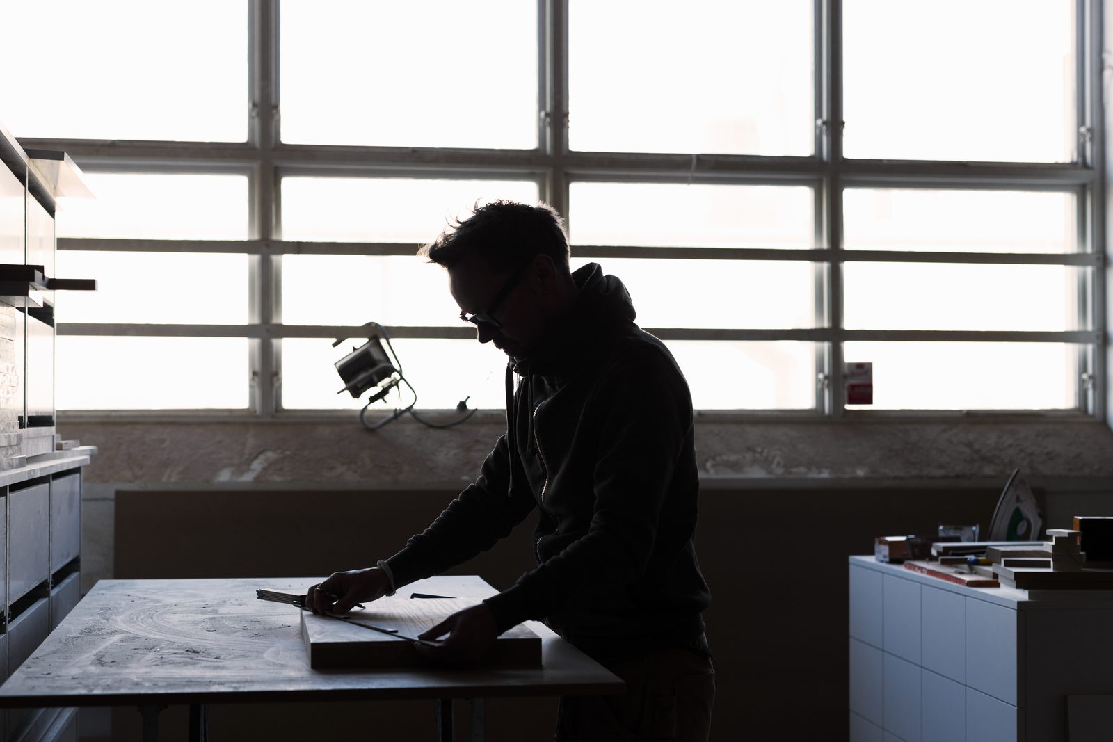 A carpenter silhouetted against a bright workshop window, measuring a board on the bench