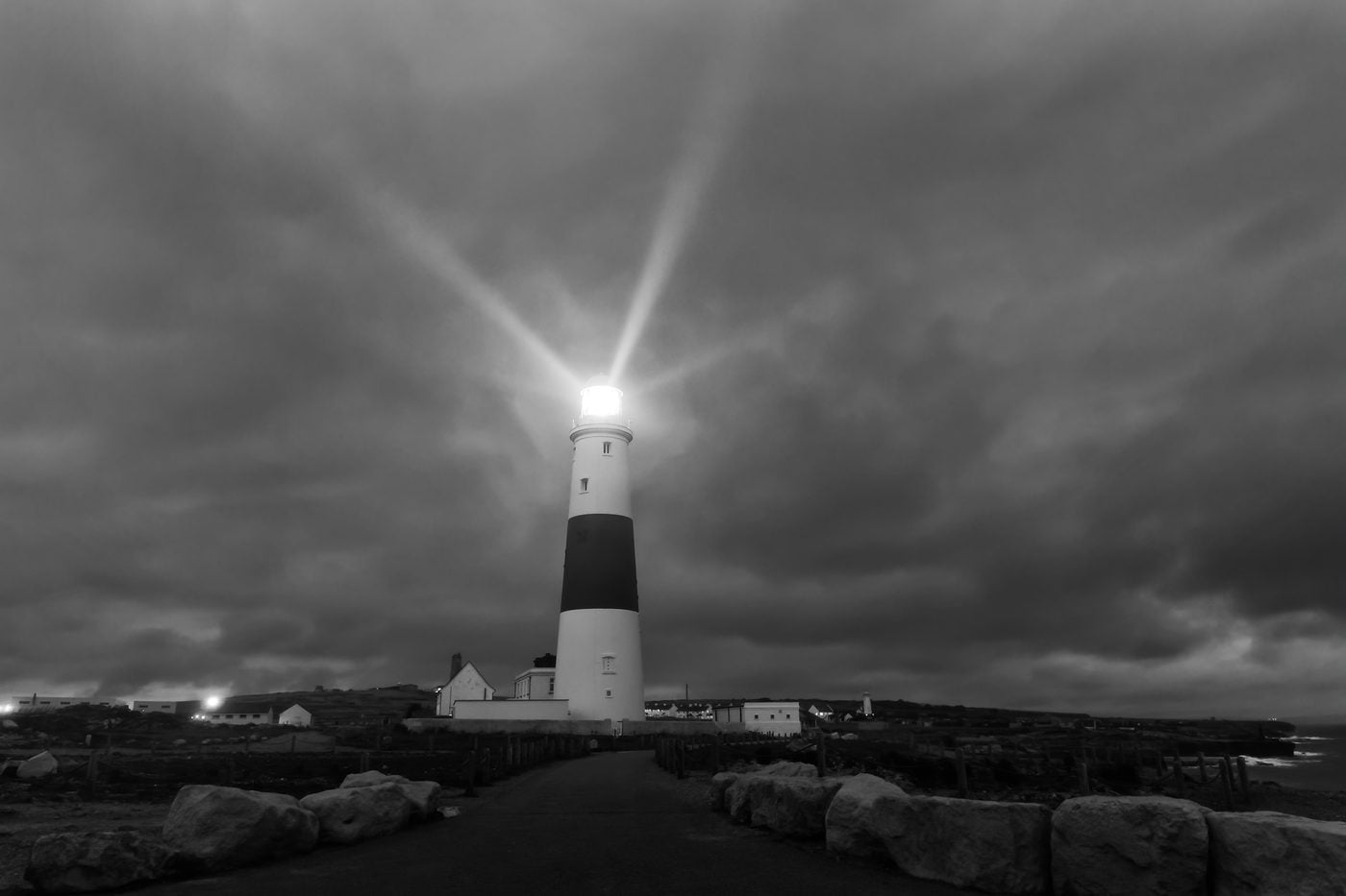 A black and white banded lighthouse on a rocky headland, its beam flaring into heavy cloud