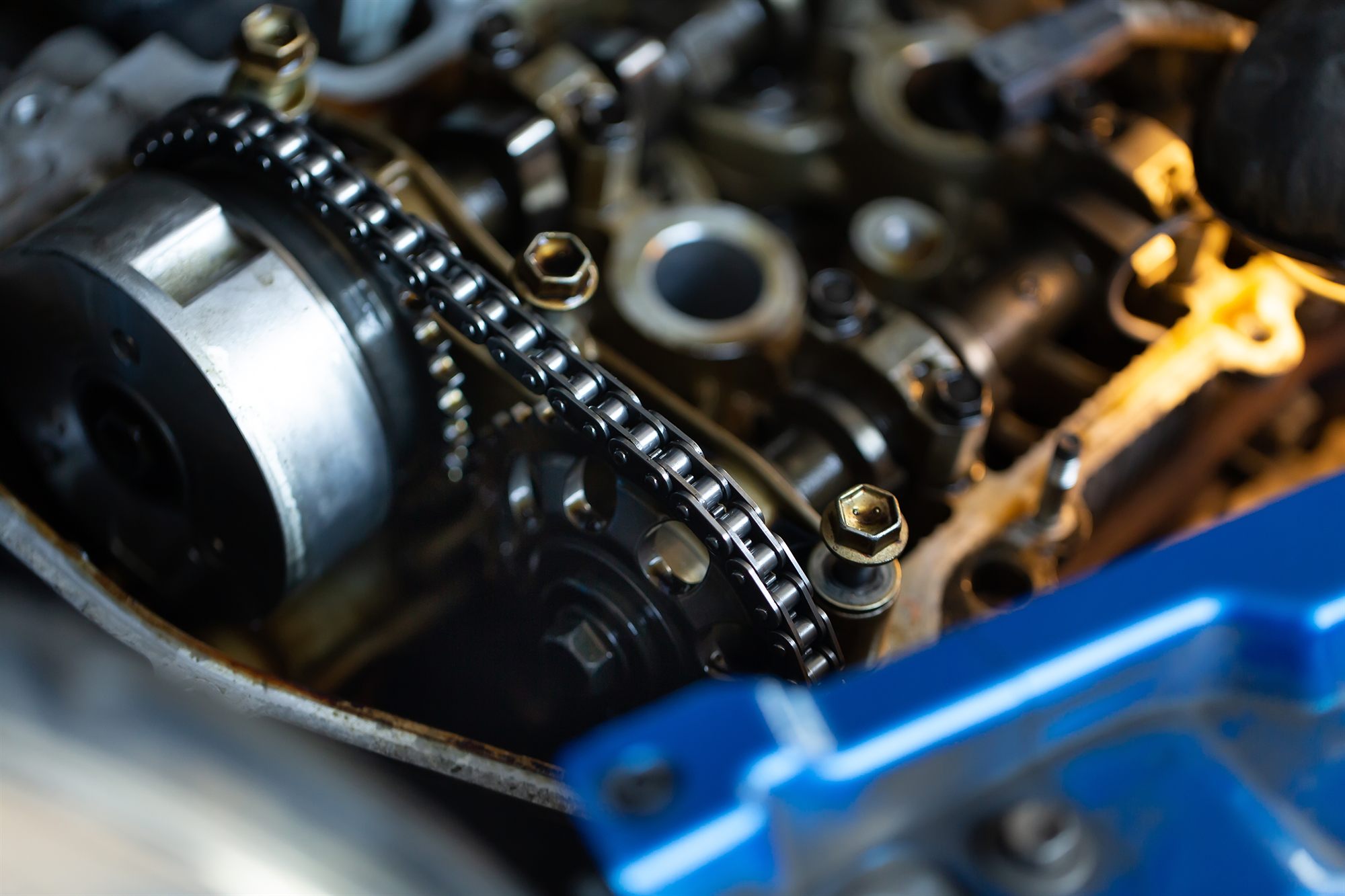 An engine opened up in close-up, the timing chain running over the cam gear past bolt heads and a blue cover